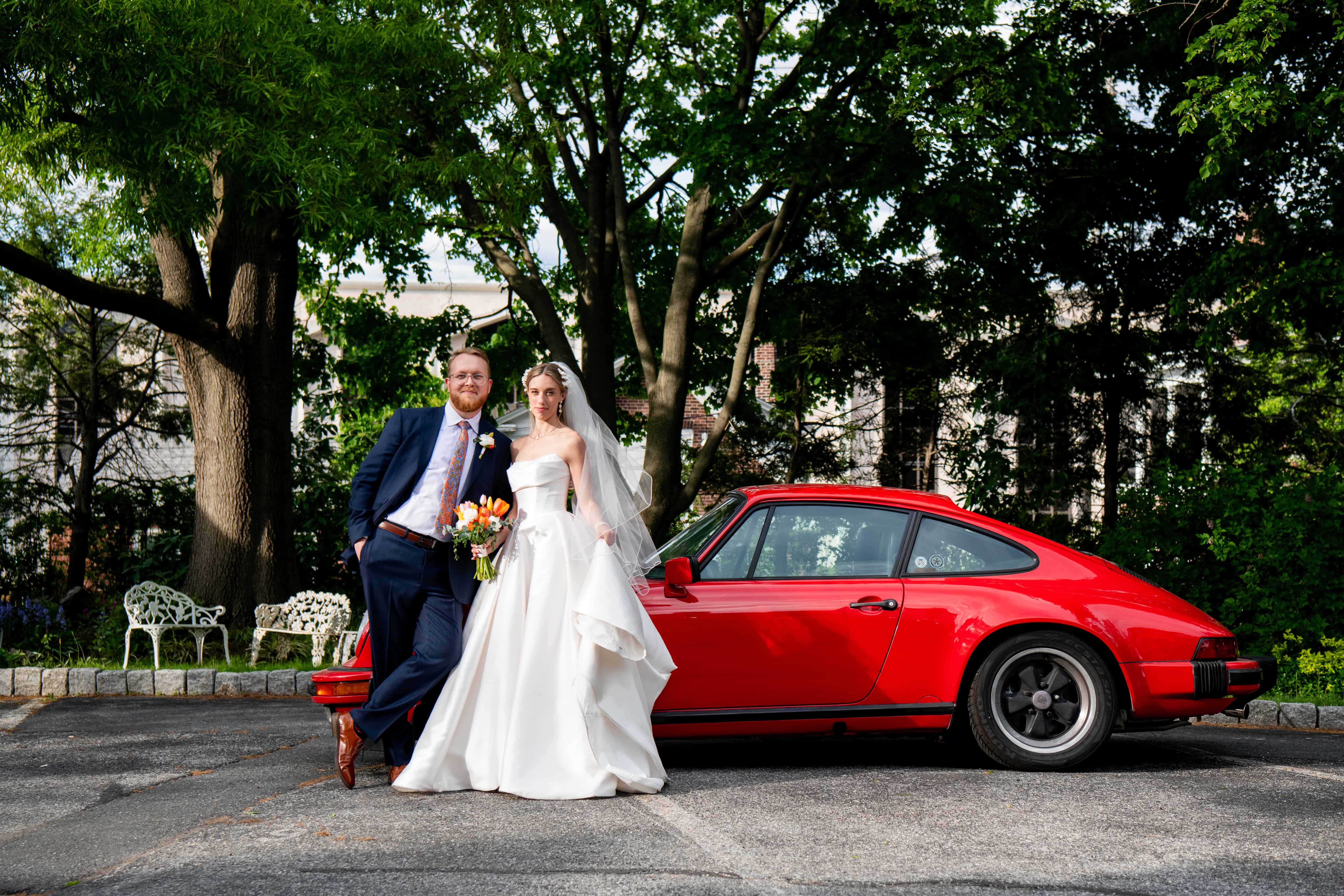 Bride in white gown and groom in blue suit pose naturally by red sports car outdoors; wedding photography inspiration.