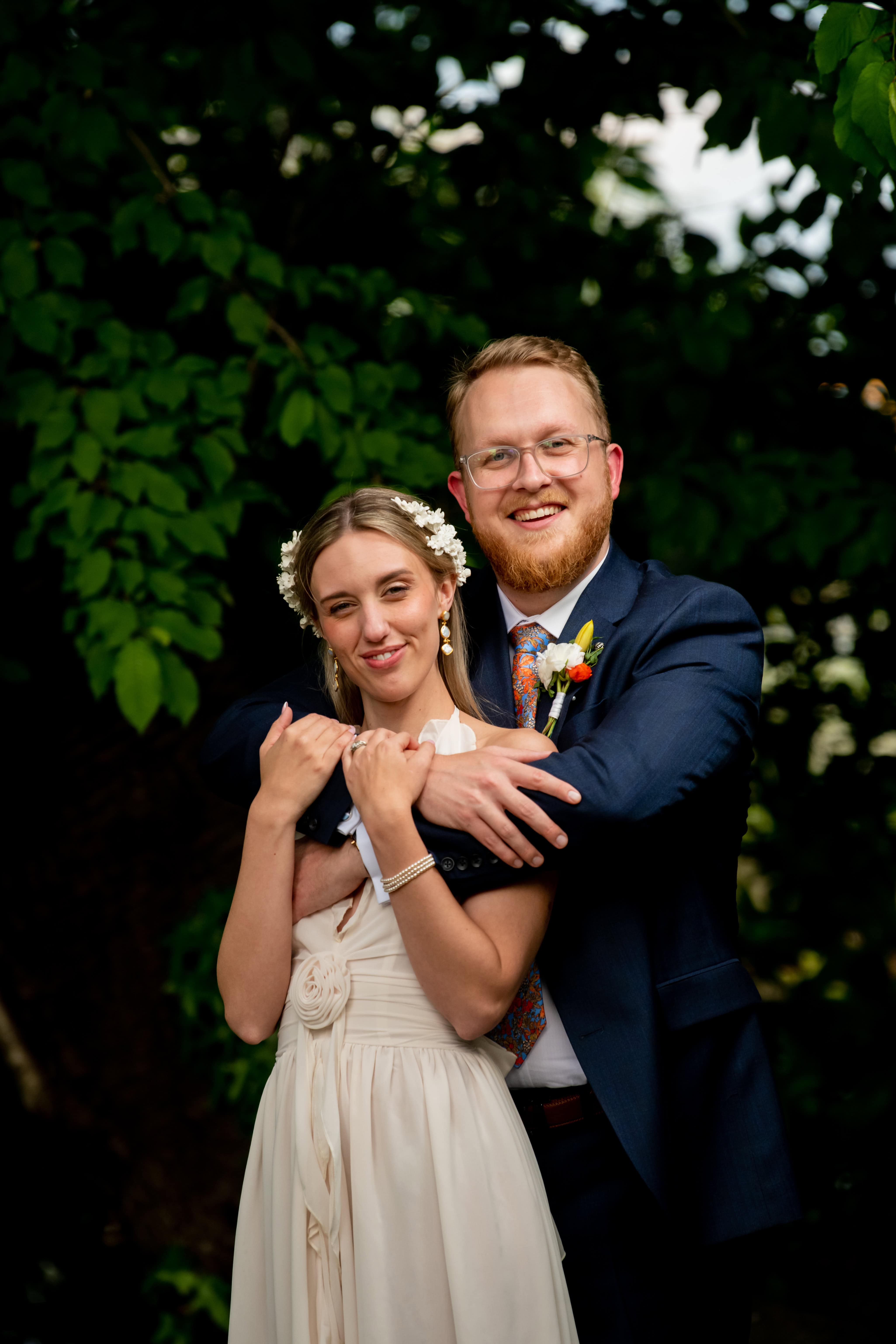 Joyful bride and groom embrace outdoors by green trees, smiling naturally in wedding photo with flowers in their hair.