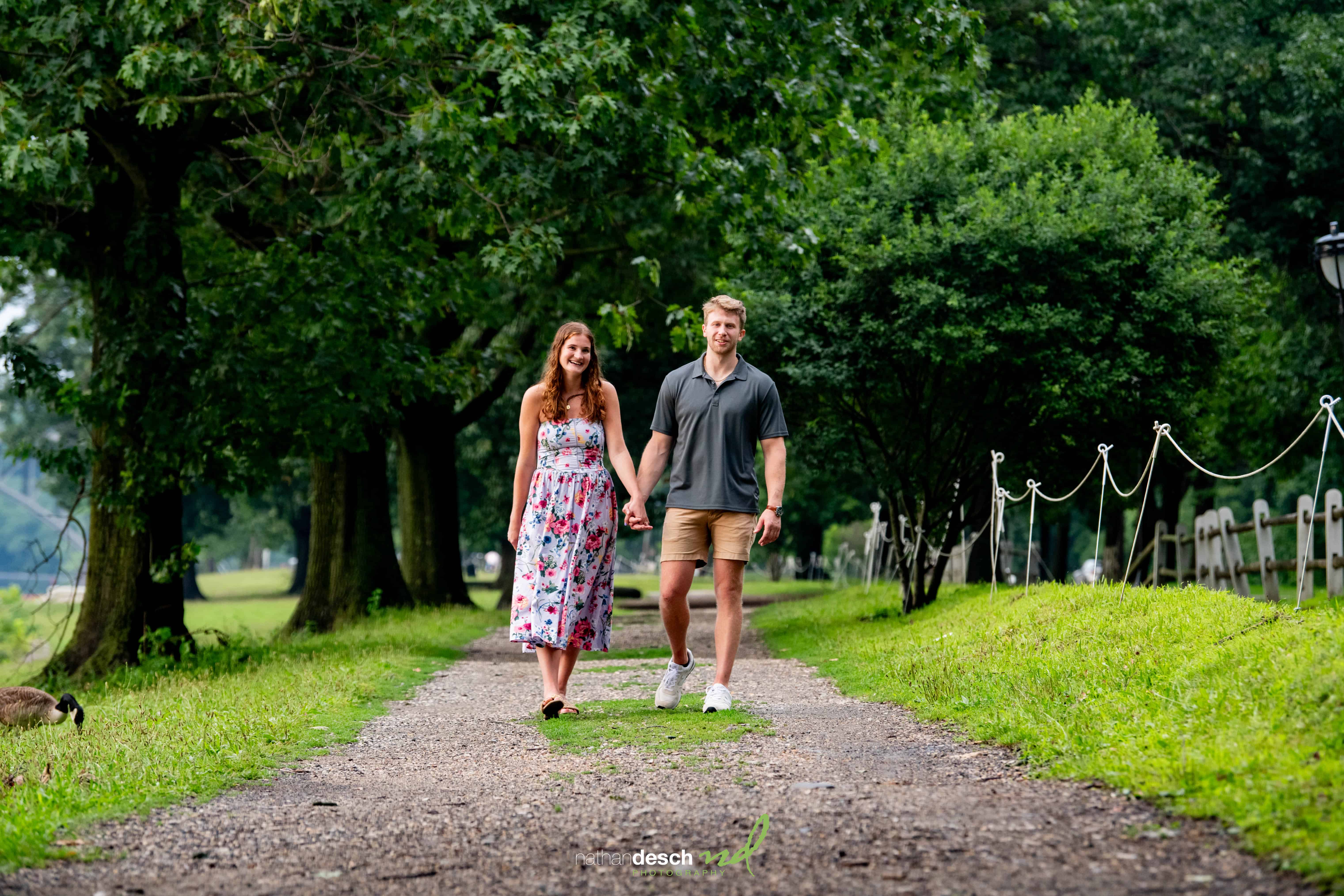 Engaged couple walking hand in hand down a sunlit, tree-lined park path, sharing smiles—romantic outdoor engagement photo.