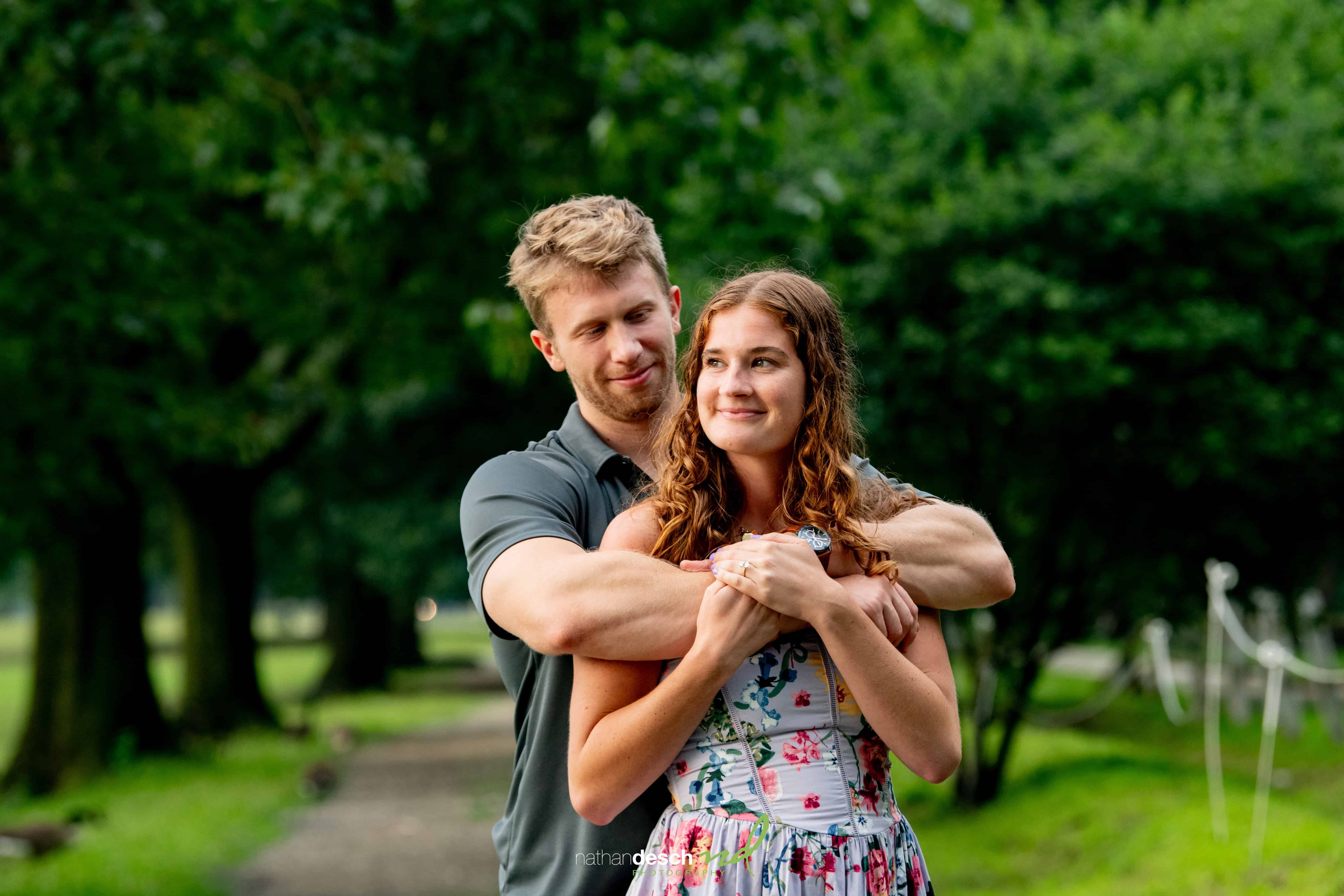 Engaged couple embracing on a scenic park path, woman in floral dress and man in dark shirt, surrounded by lush greenery.