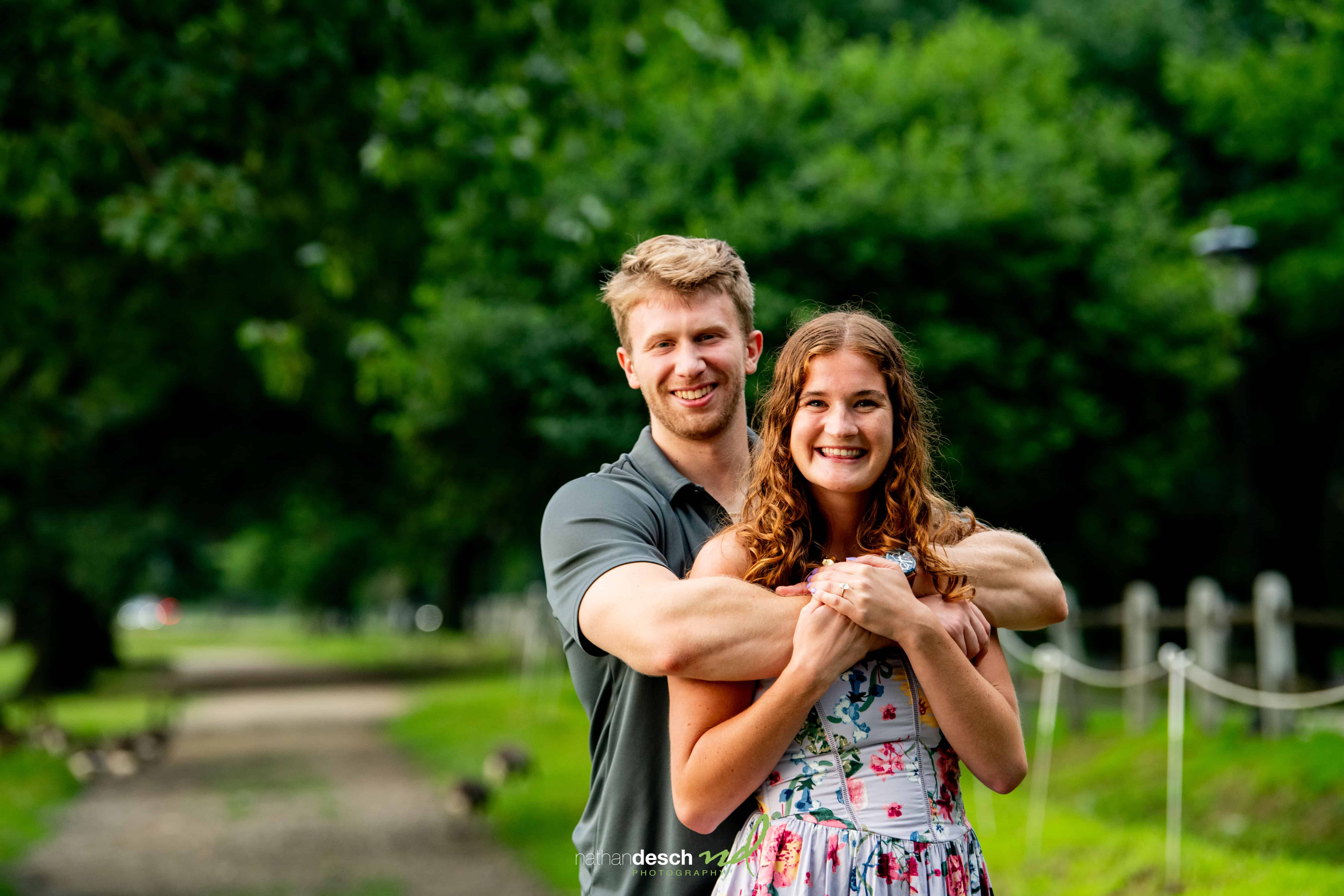 Happy couple embracing on a tree-lined path, groom in gray shirt hugs bride in floral dress, smiling at camera.