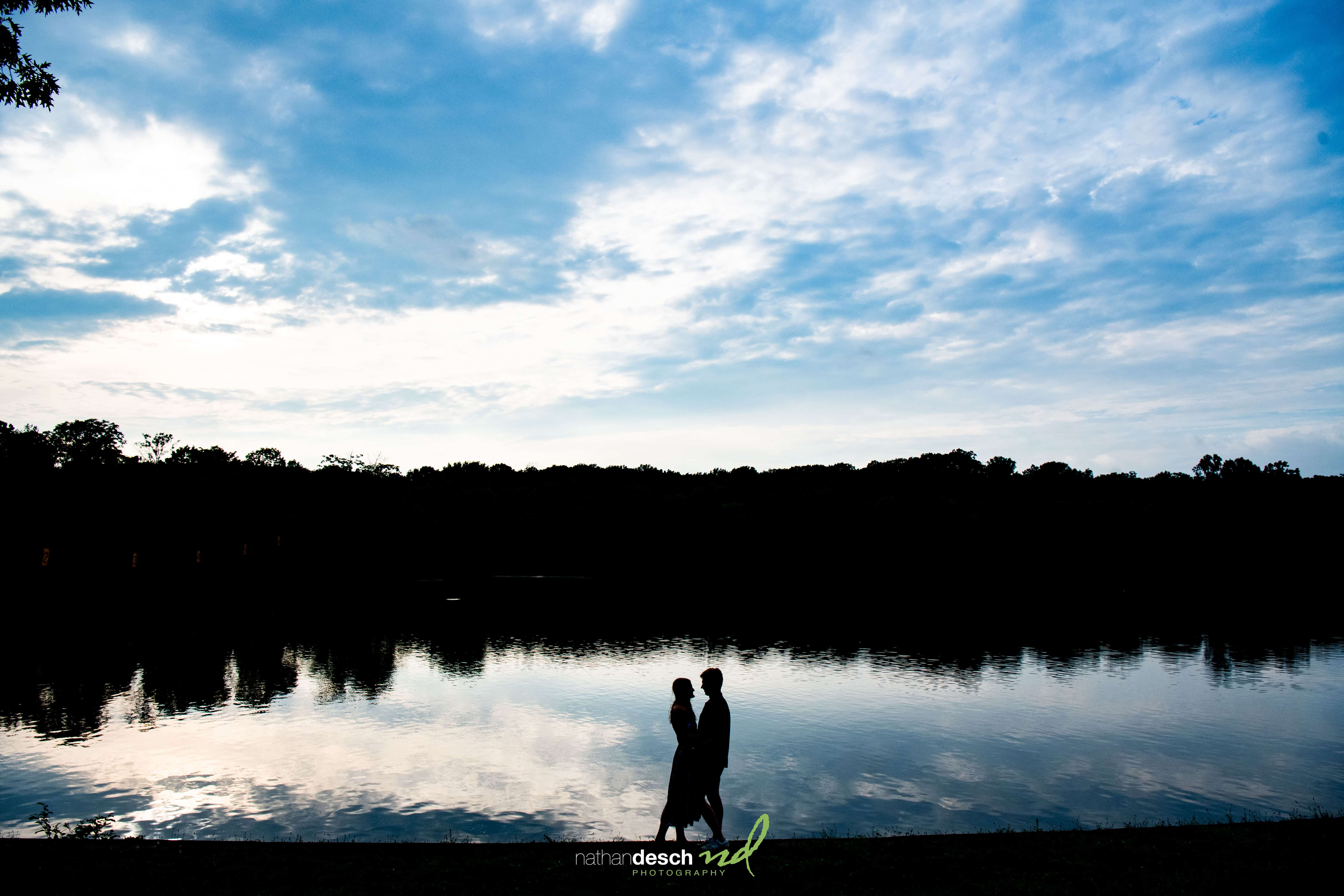 Romantic couple silhouette by a serene lake at sunset, wedding photography with trees and clouds reflected in the water.
