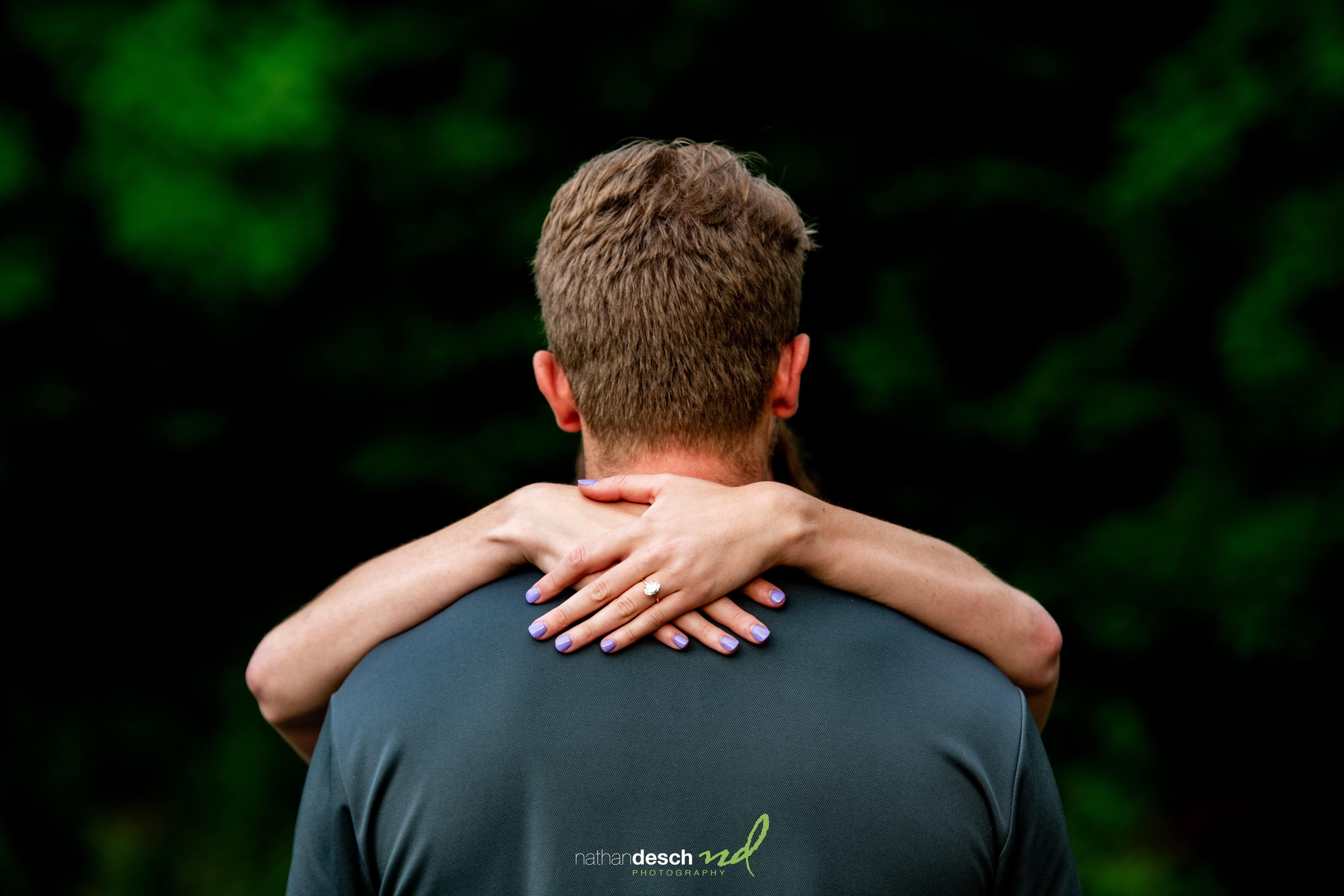 Groom embraces bride from behind, wedding ring visible on hand, set against a blurred green outdoor background.