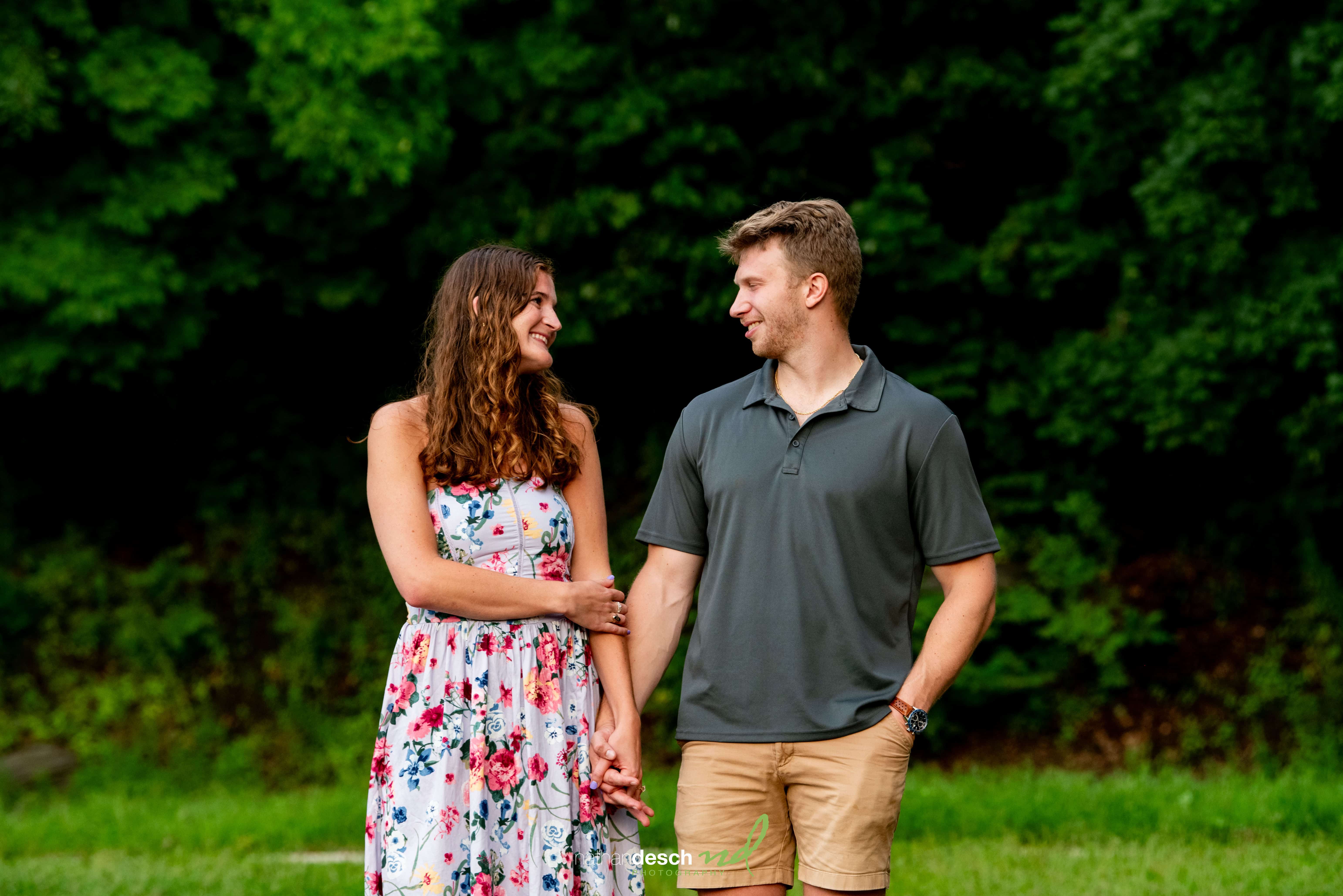 Engaged couple holding hands and smiling outdoors among lush green trees, candid wedding photography session.