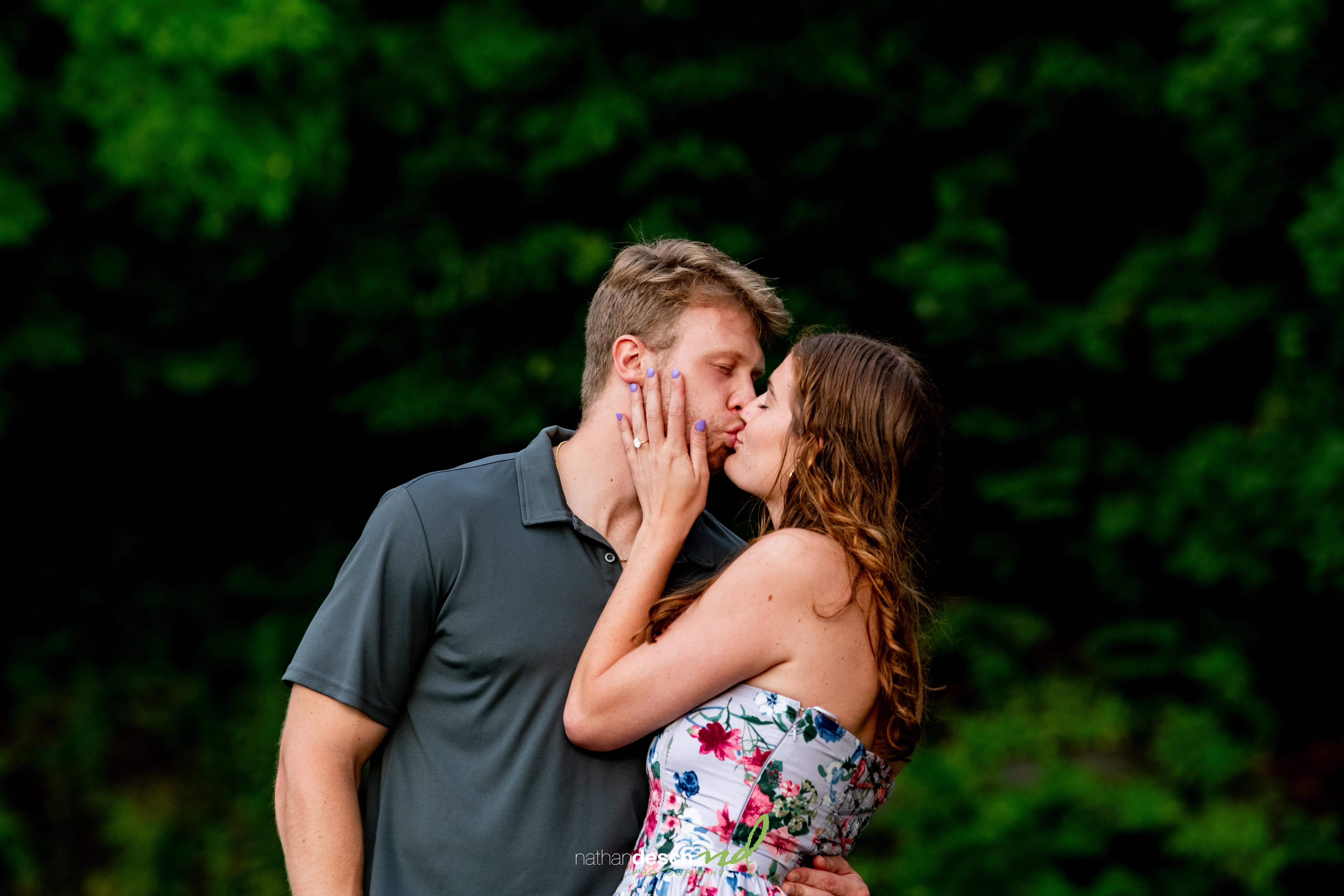 Romantic outdoor kiss between couple; woman in floral dress holds partner’s face, lush green trees softly blurred behind them.