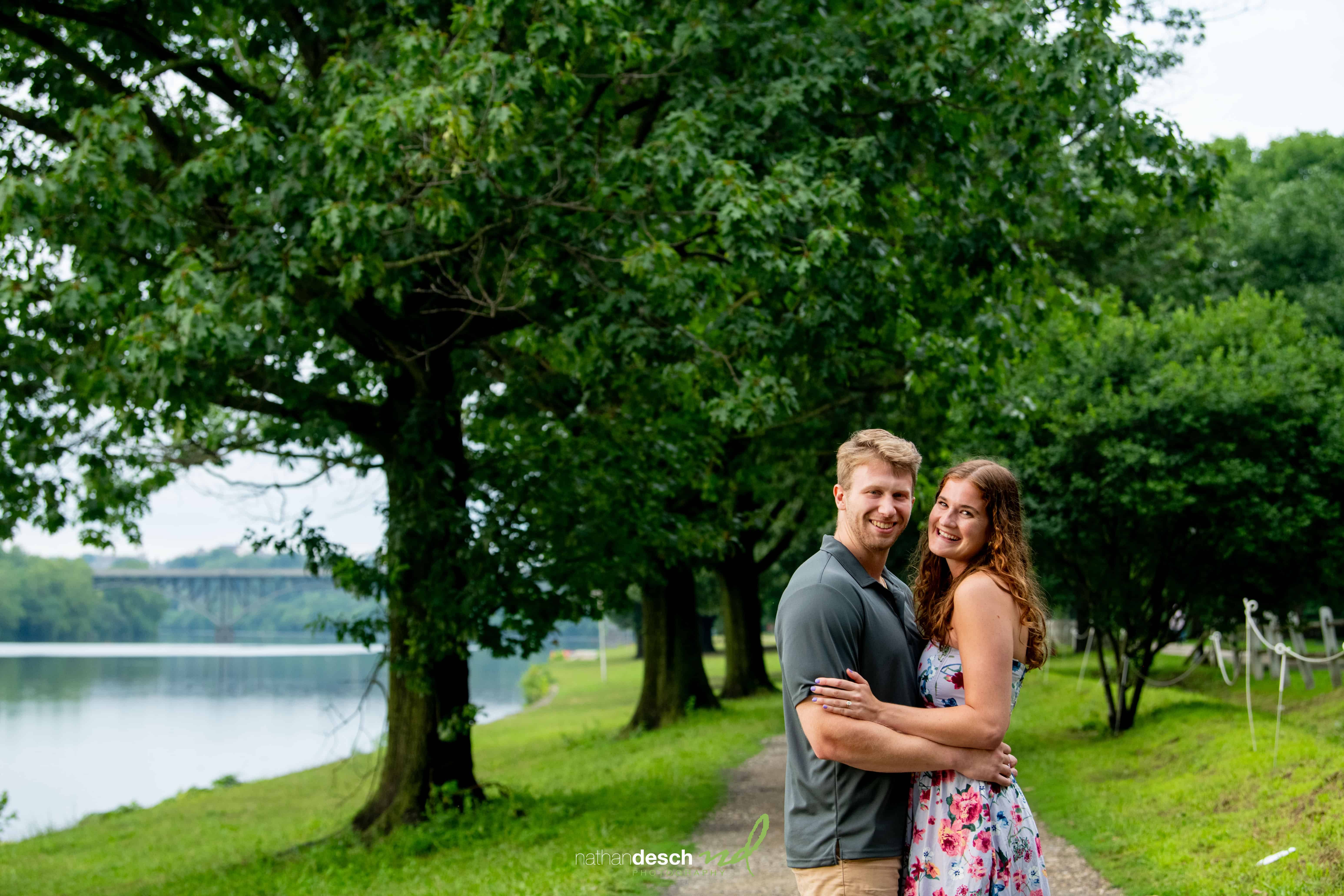Smiling couple embraces on a scenic, tree-lined riverside path with lush greenery and a distant bridge—romantic wedding photo.