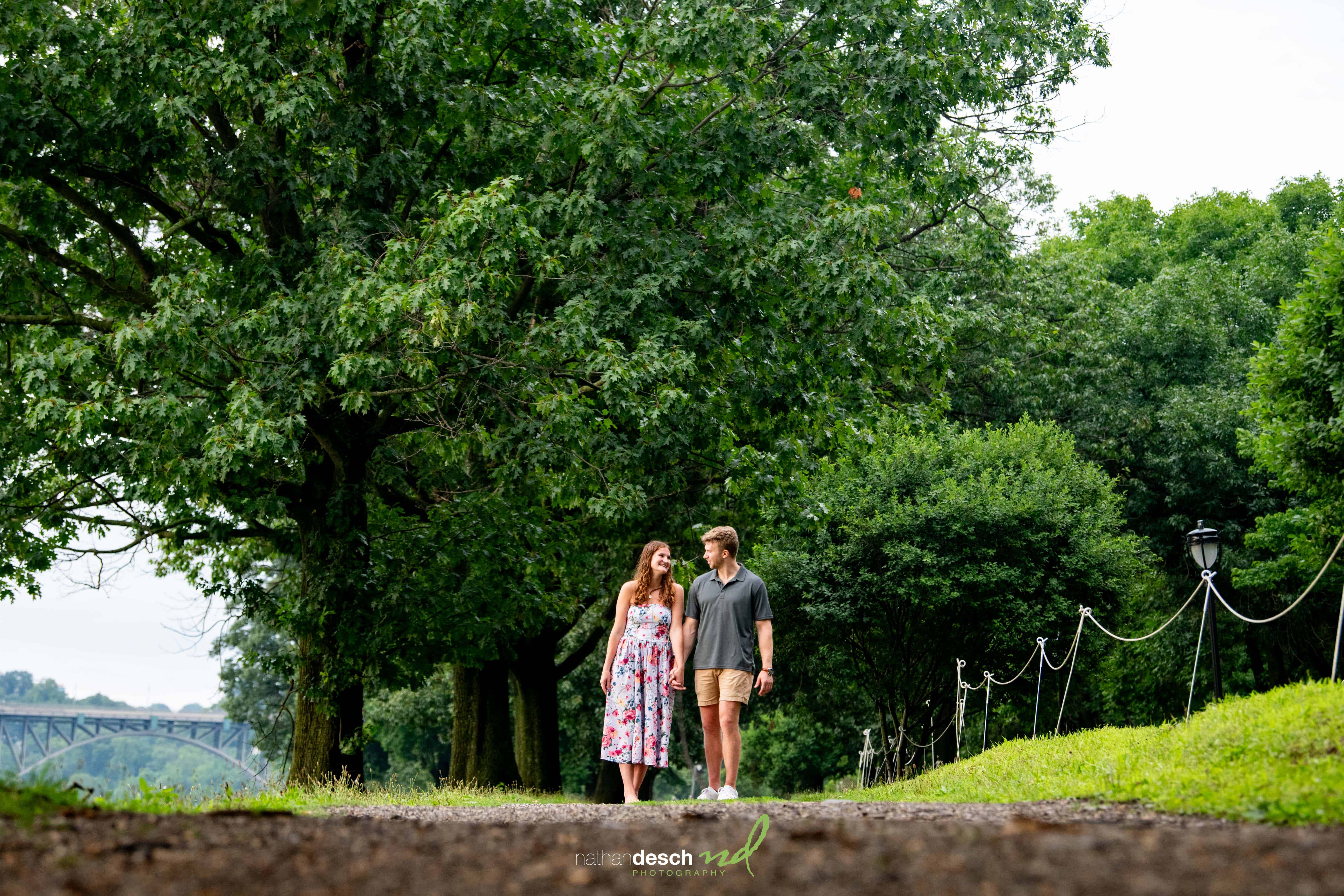 Bride and groom walk hand in hand on a scenic path lined with green trees, bridge in background, romantic outdoor wedding moment.