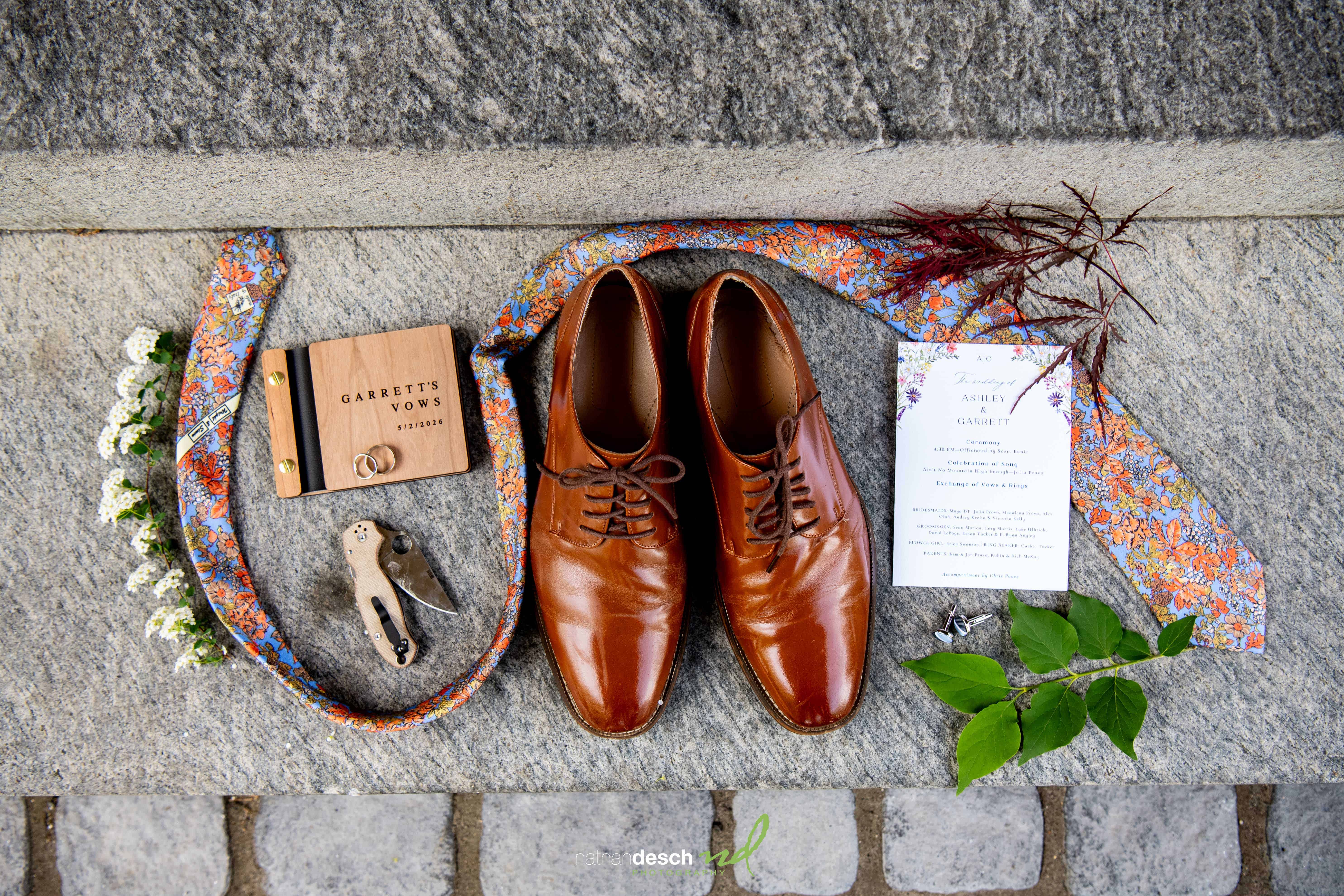Flat lay of brown dress shoes, floral tie, rings, boutonniere, vows book, and invites for University and Whist Club wedding.