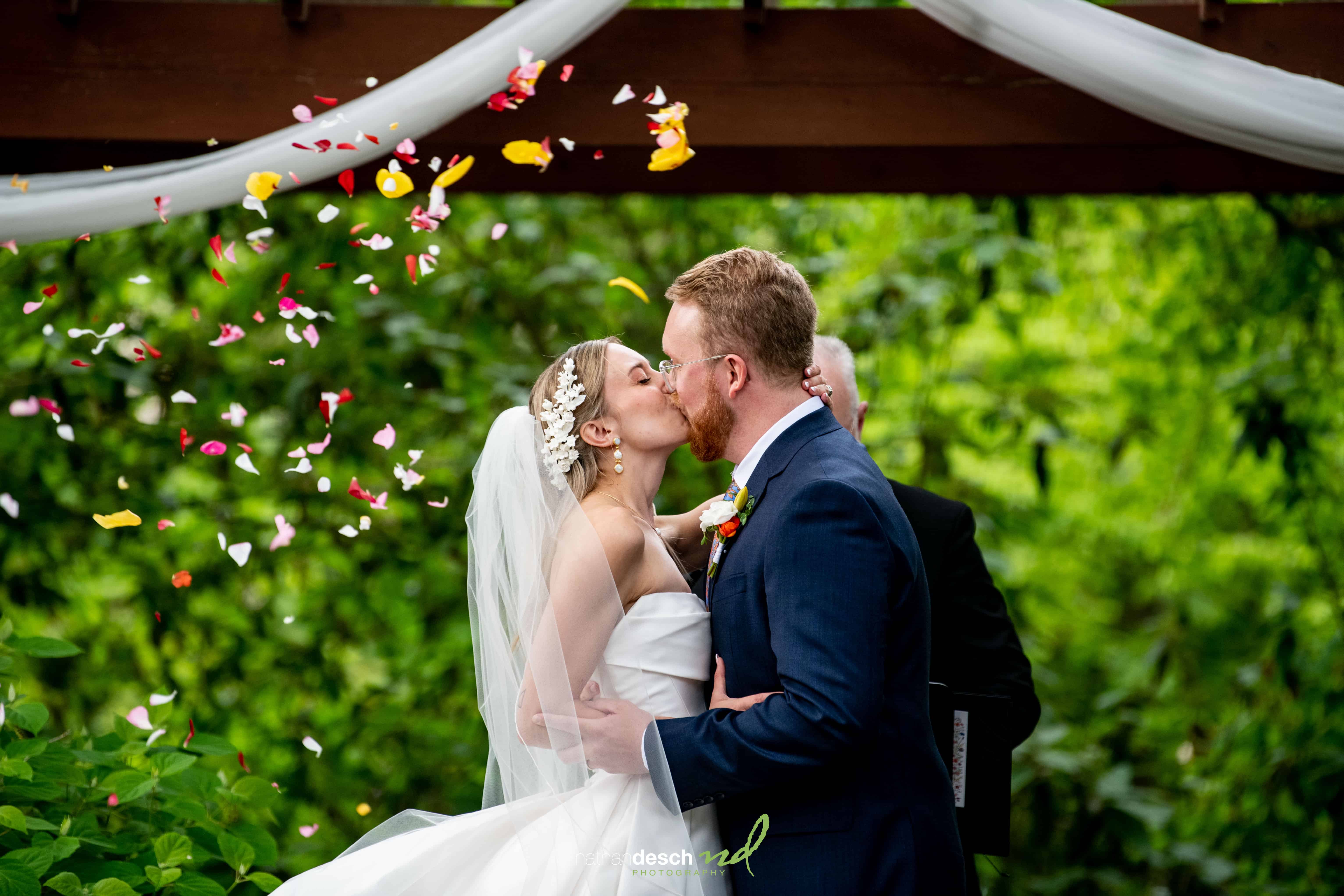 Bride and groom kiss under a pergola amid greenery with flower petals tossed at their University and Whist Club wedding.