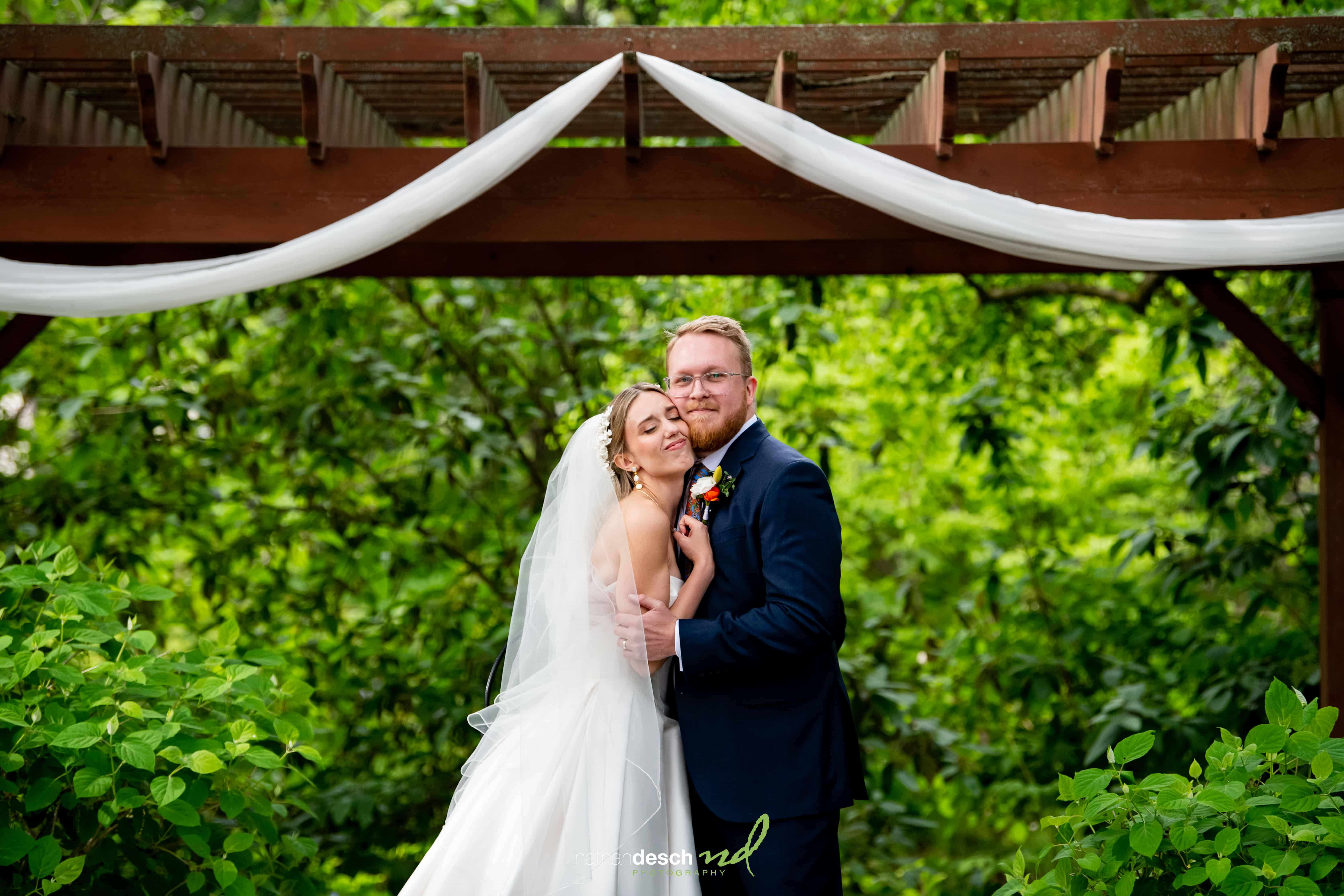 Bride and groom embrace under a white-draped arbor with greenery at their University and Whist Club wedding.