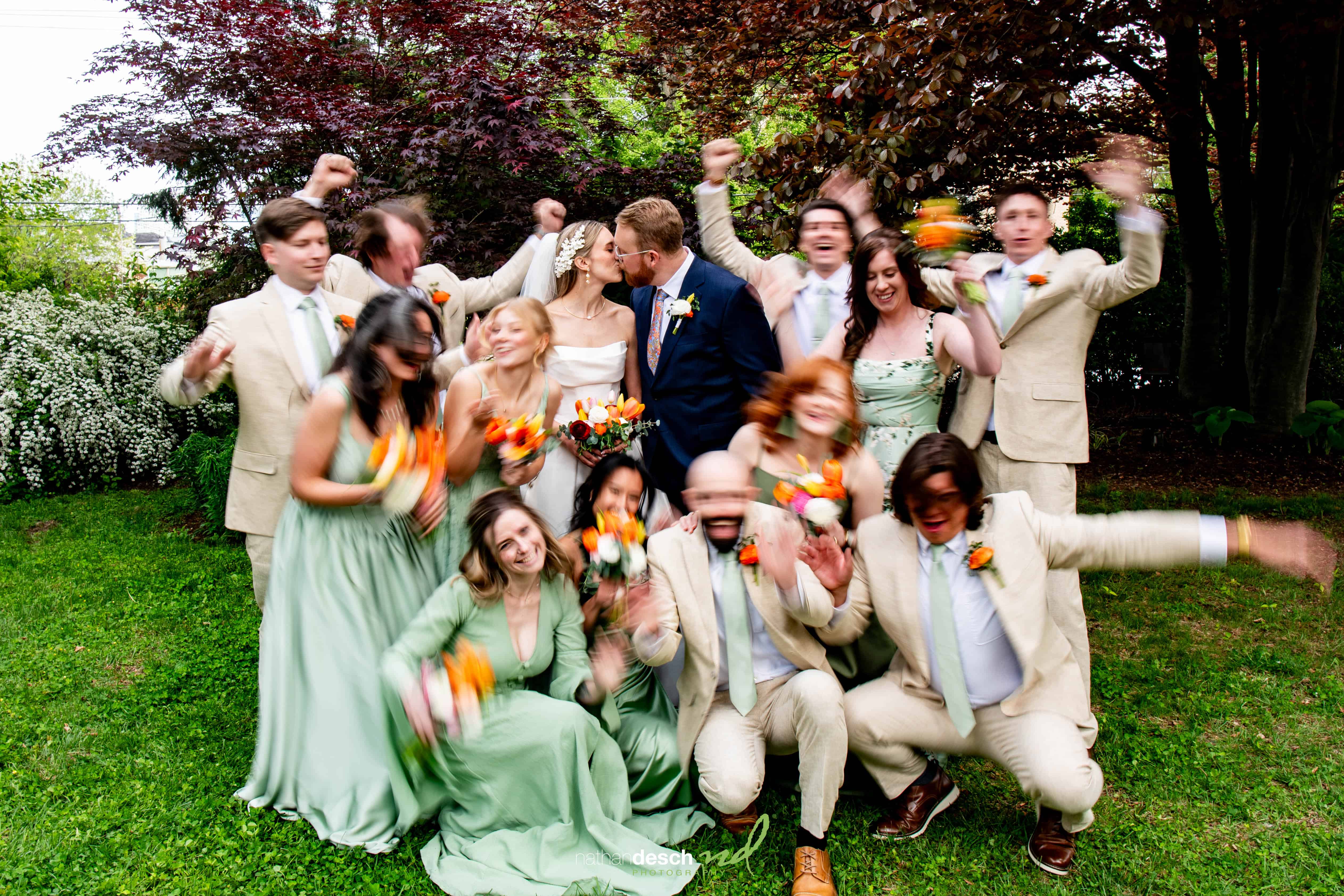 Bride and groom kiss as wedding party in green dresses and light suits cheer outdoors at University and Whist Club wedding.