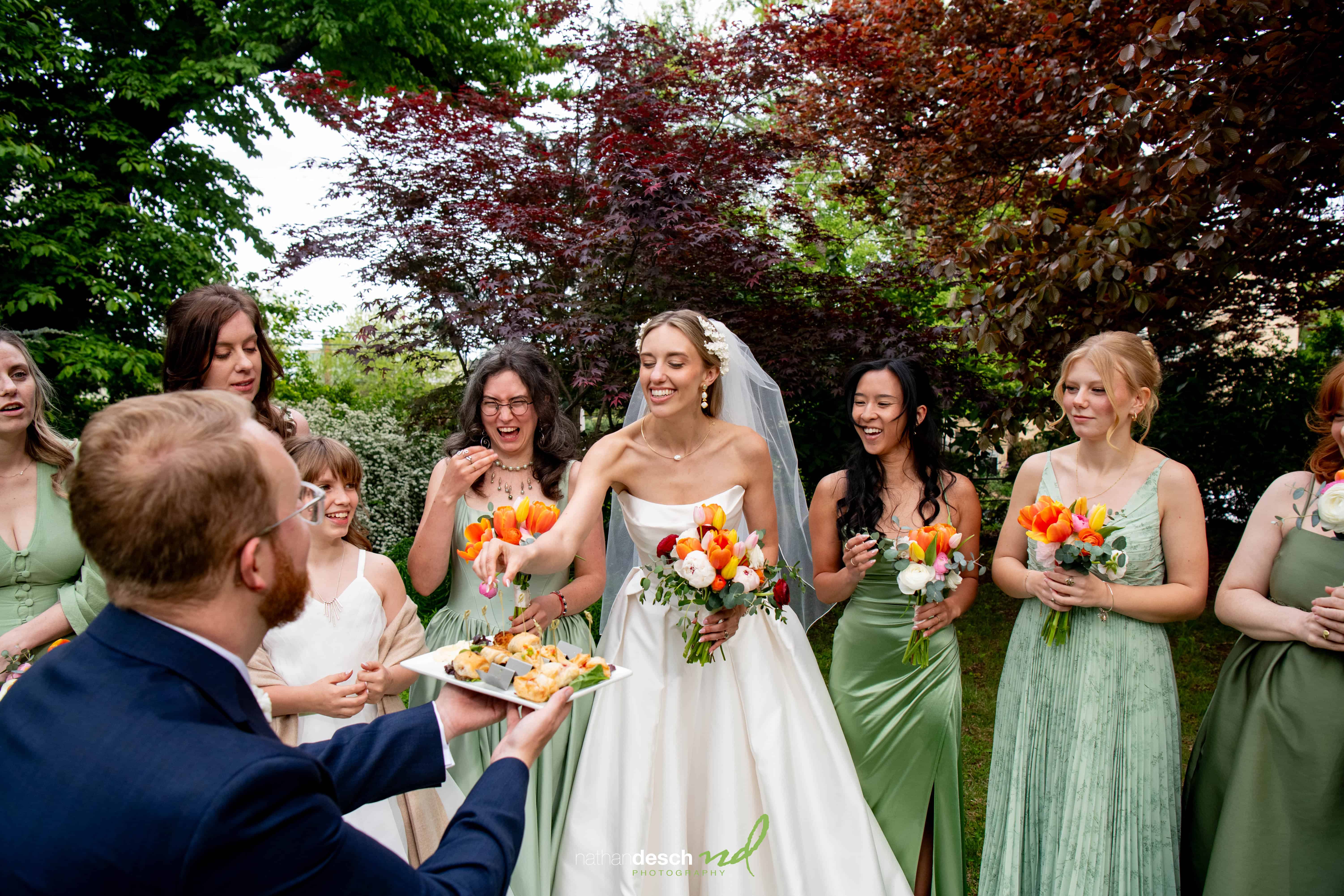 Bride in white dress smiles at University and Whist Club wedding, surrounded by bridesmaids in green with orange bouquets outdoors.