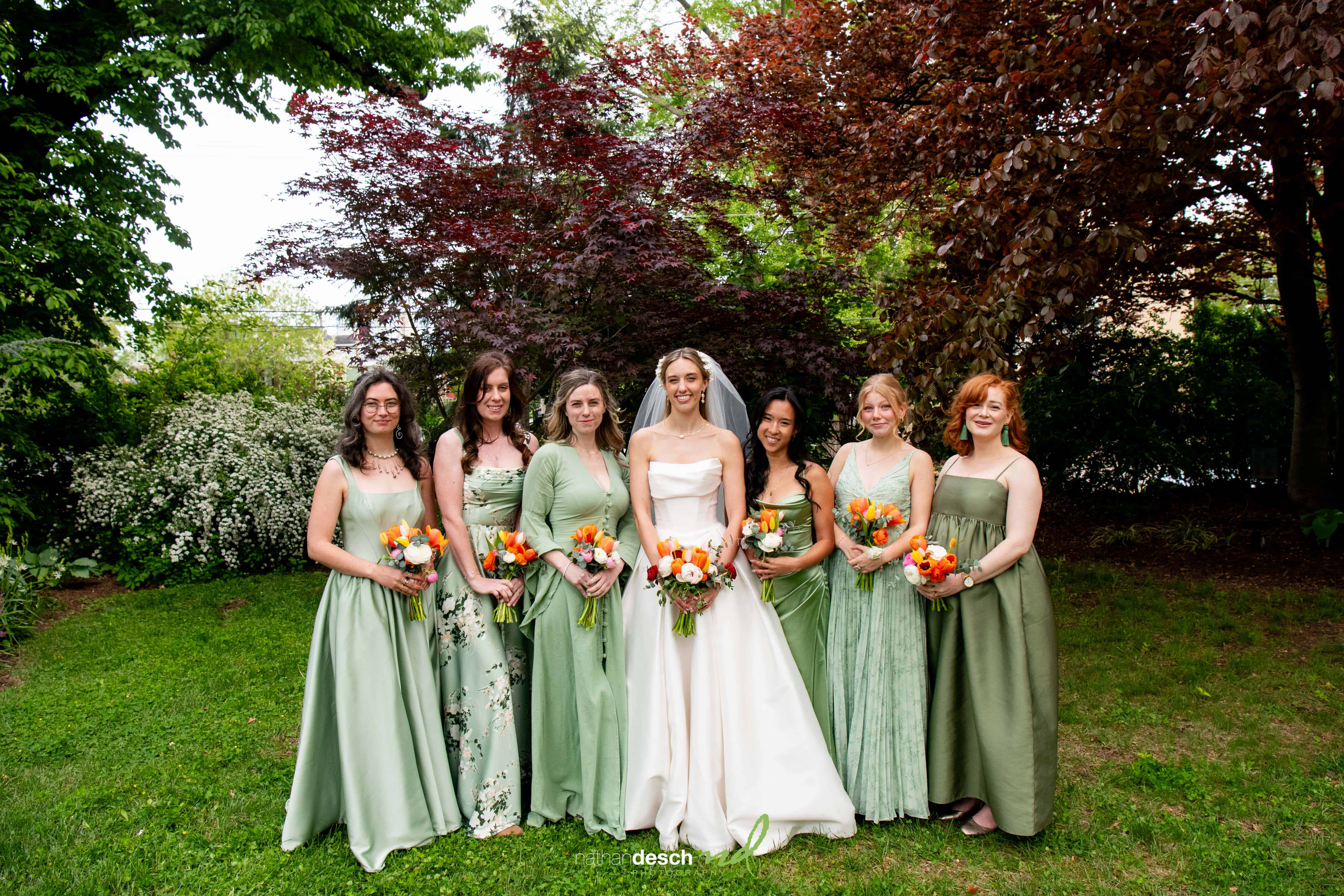 Bride and six bridesmaids in green dresses pose with orange bouquets in lush garden at University and Whist Club wedding.