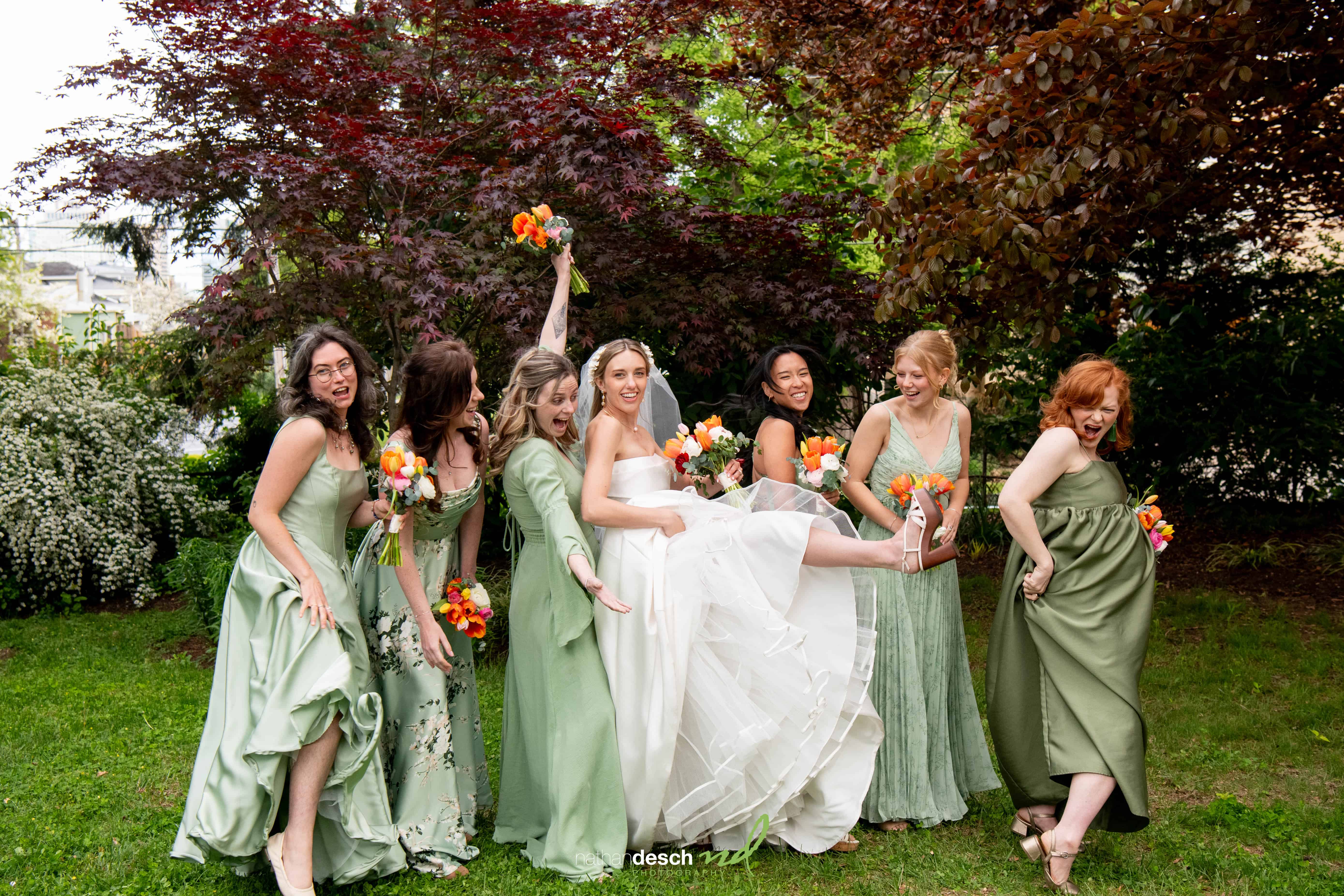 Bride in white dress and six bridesmaids in green dresses laughing with bouquets at University and Whist Club garden wedding.