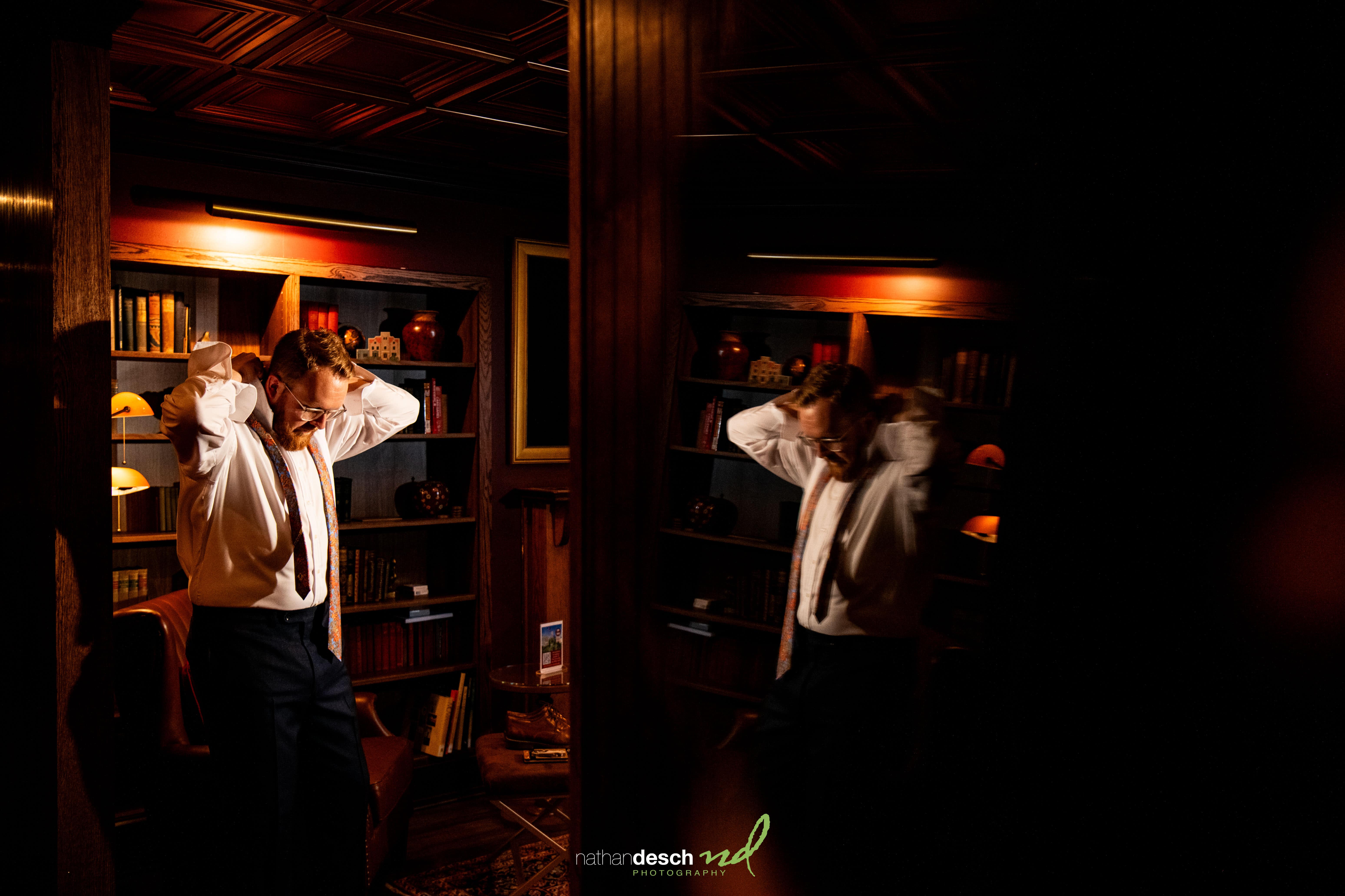 Groom adjusts tie in warmly lit University and Whist Club library, ready for Garrett and Ashley's wedding, mirrored reflection visible.