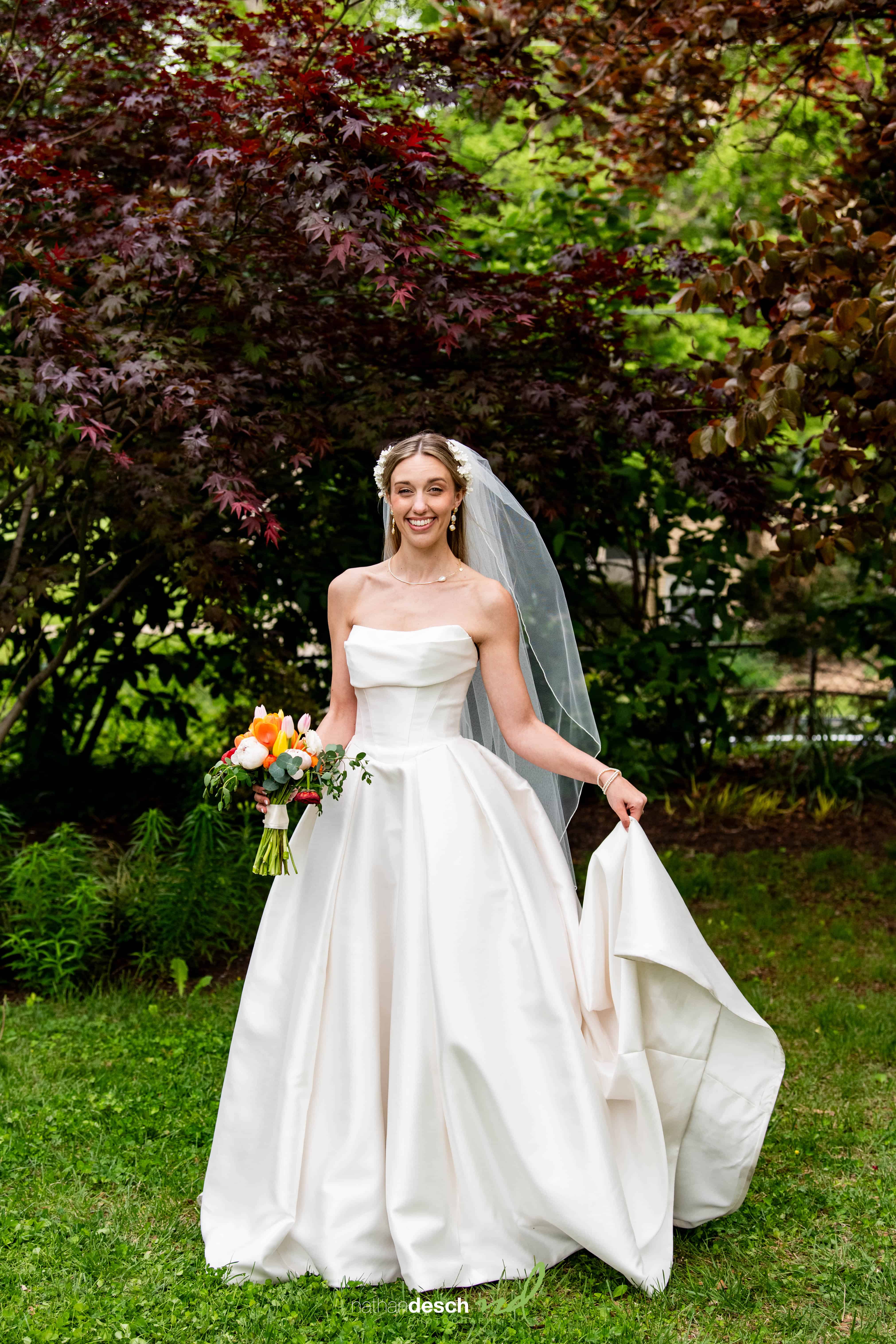 Smiling bride in strapless white gown and veil with bouquet on lawn, greenery behind at University and Whist Club wedding.