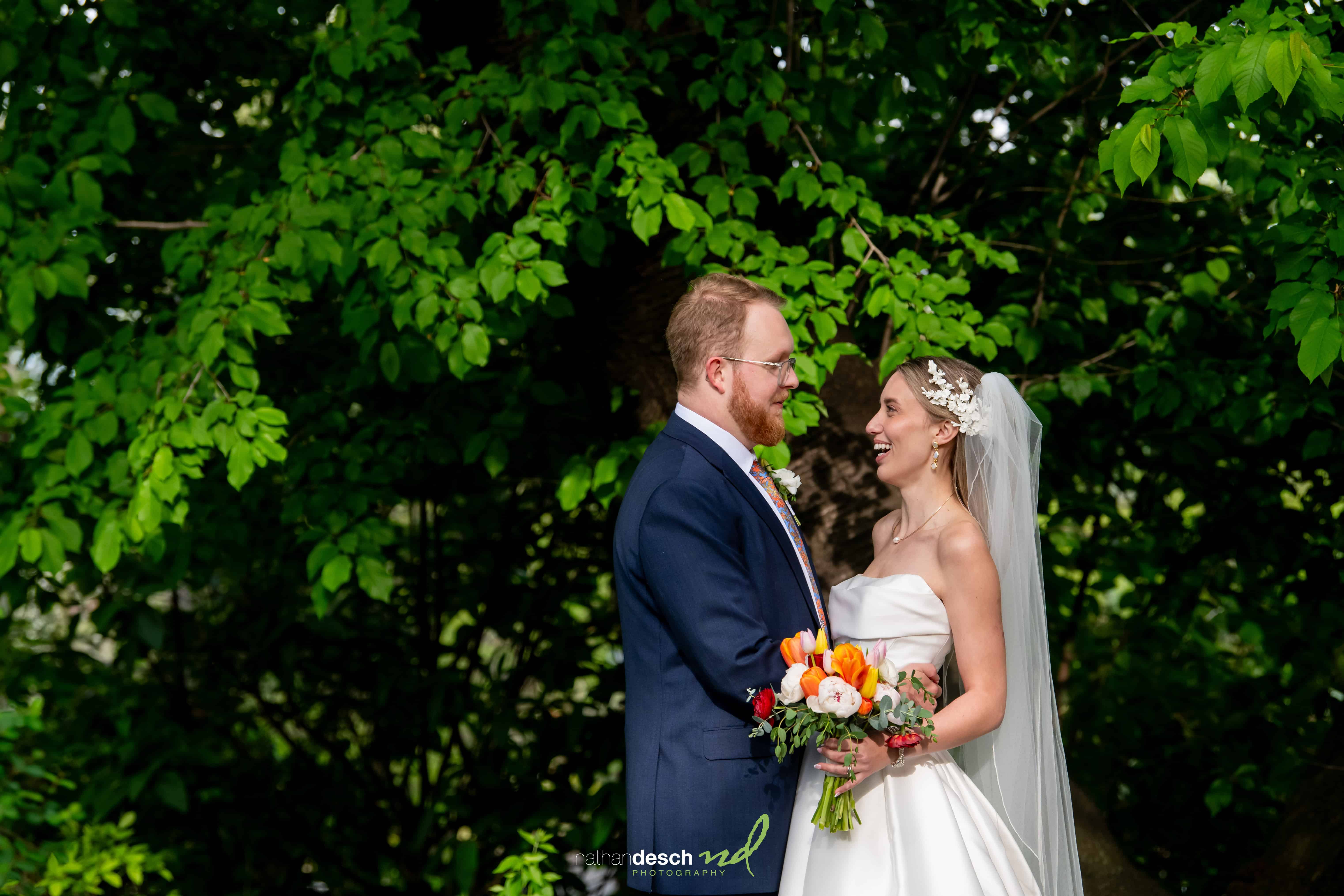 Bride and groom smile outdoors on wedding day at University and Whist Club, bride in white dress holding bouquet, lush greenery behind.