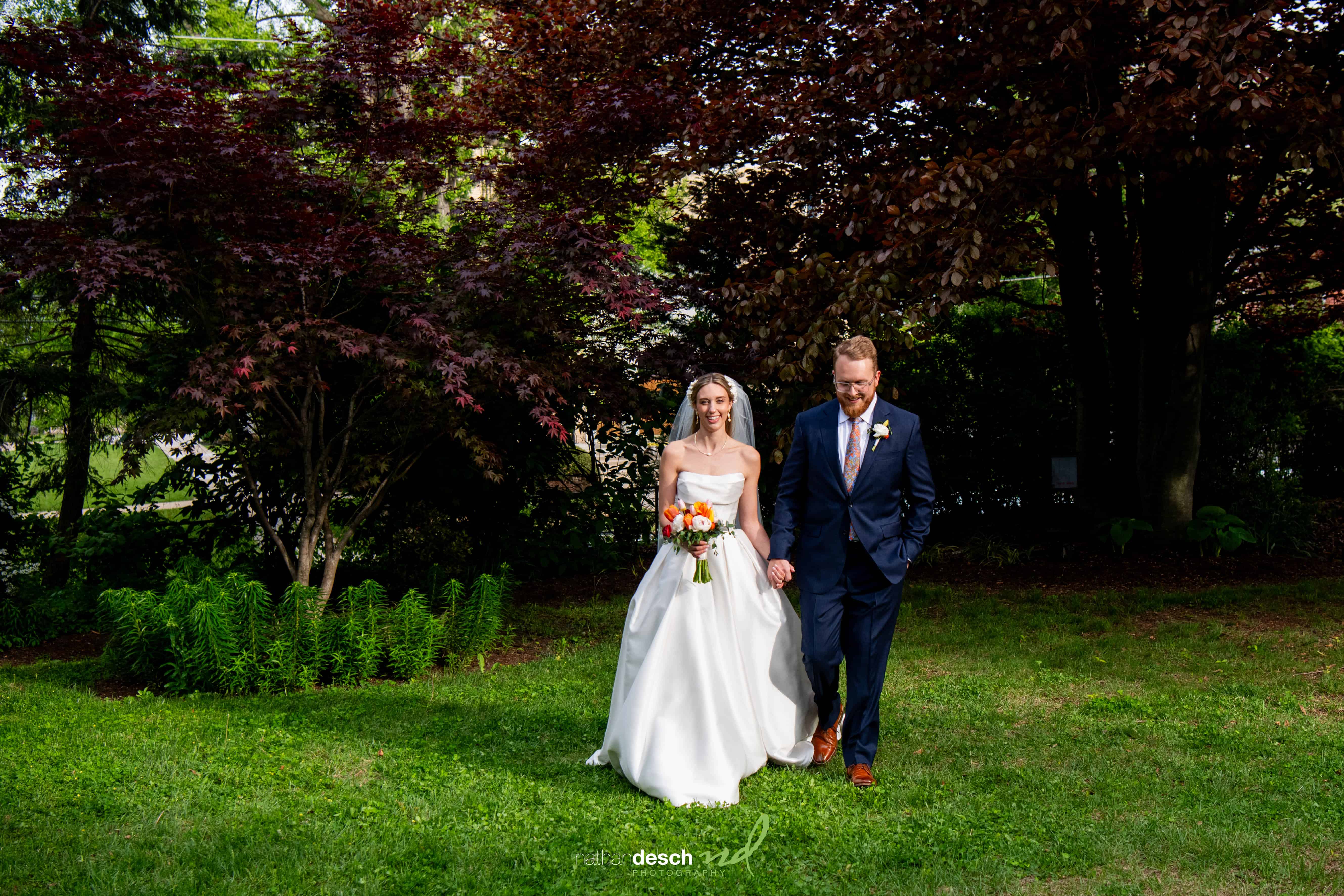 Bride and groom holding hands outdoors at University and Whist Club wedding, smiling amid green grass and colorful trees.