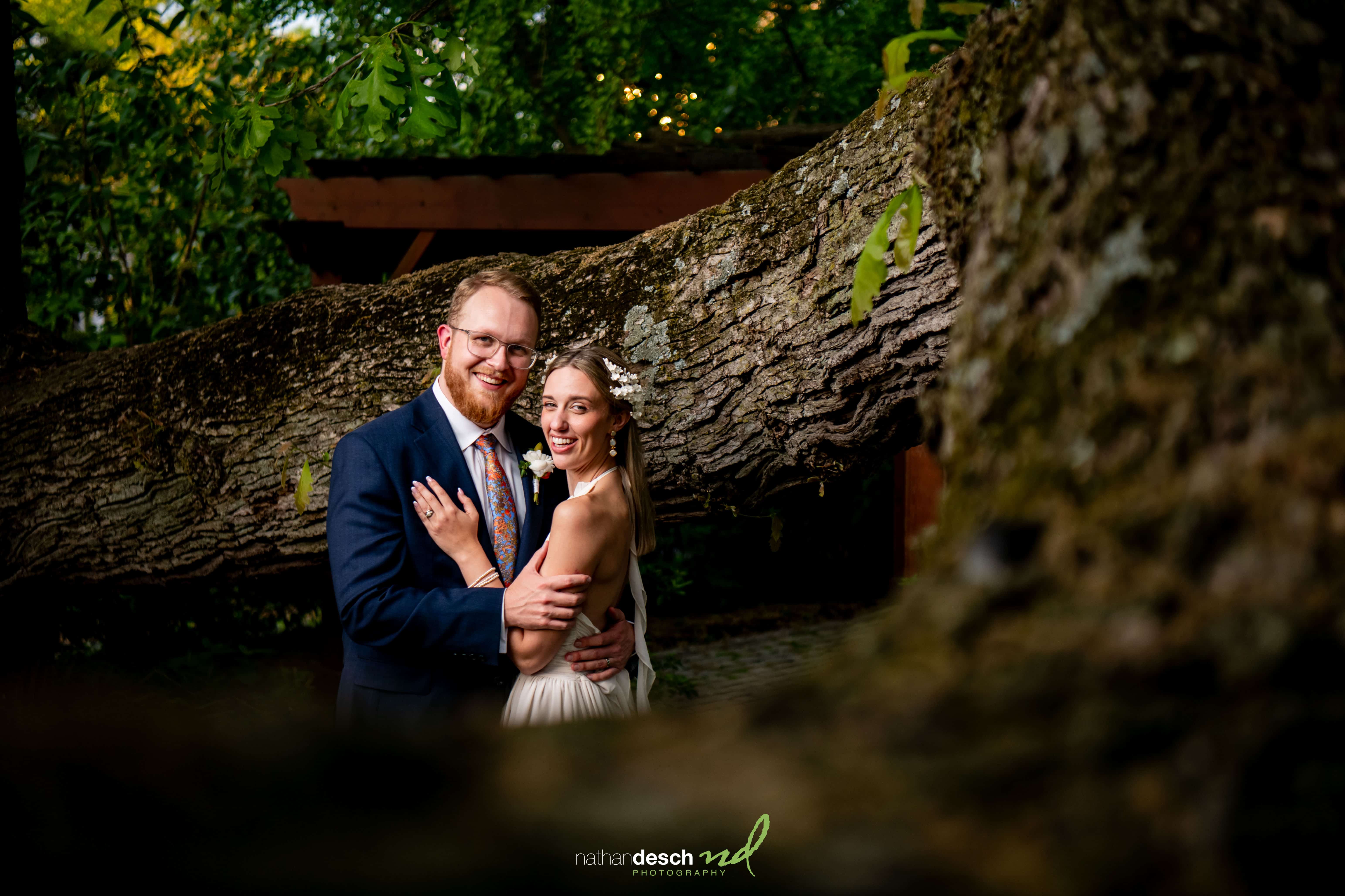 Bride and groom smiling and embracing under a fallen tree branch at University and Whist Club wedding, lush greenery around.