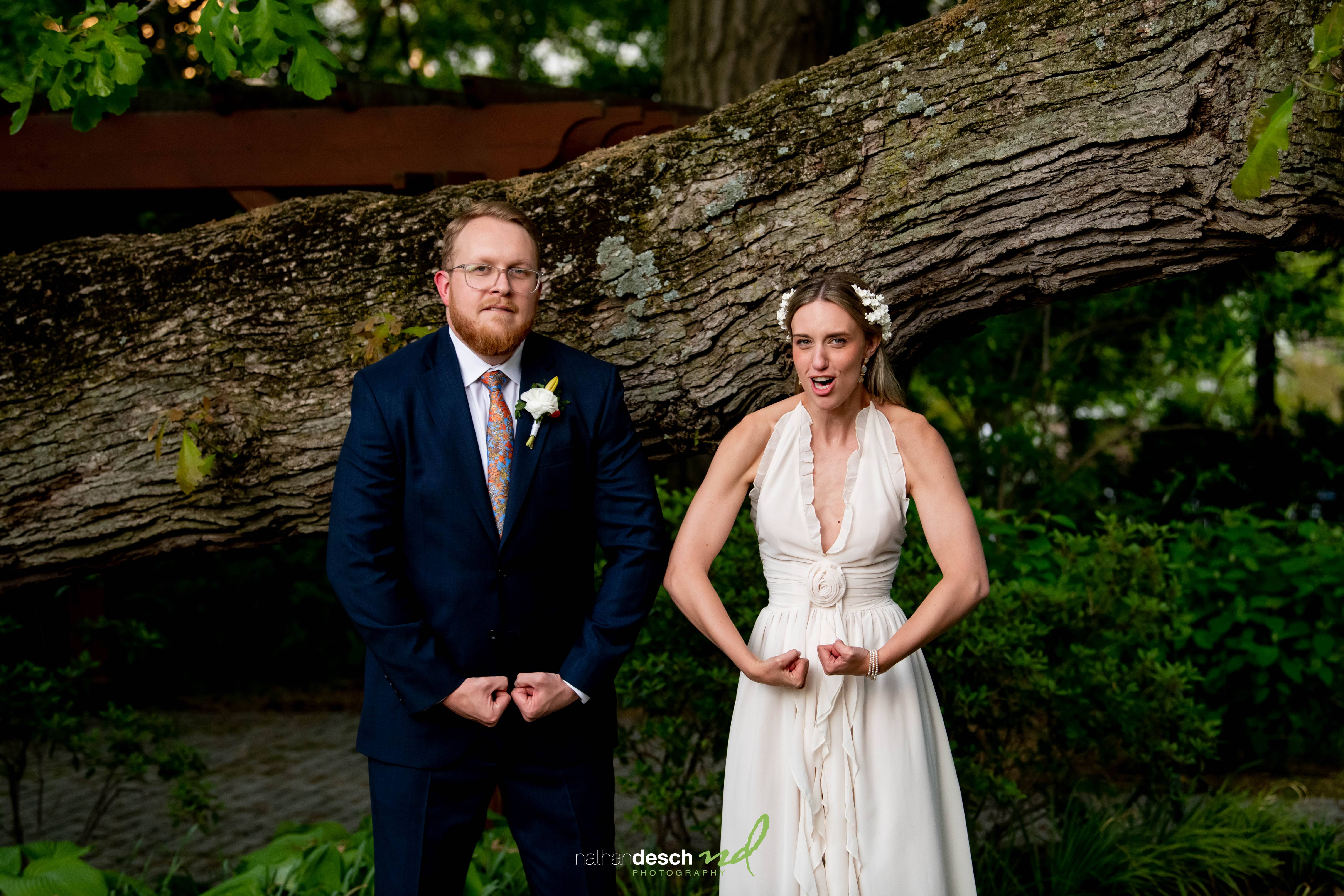 Bride and groom in wedding attire at University and Whist Club, flexing arms and making silly faces by a large tree branch.