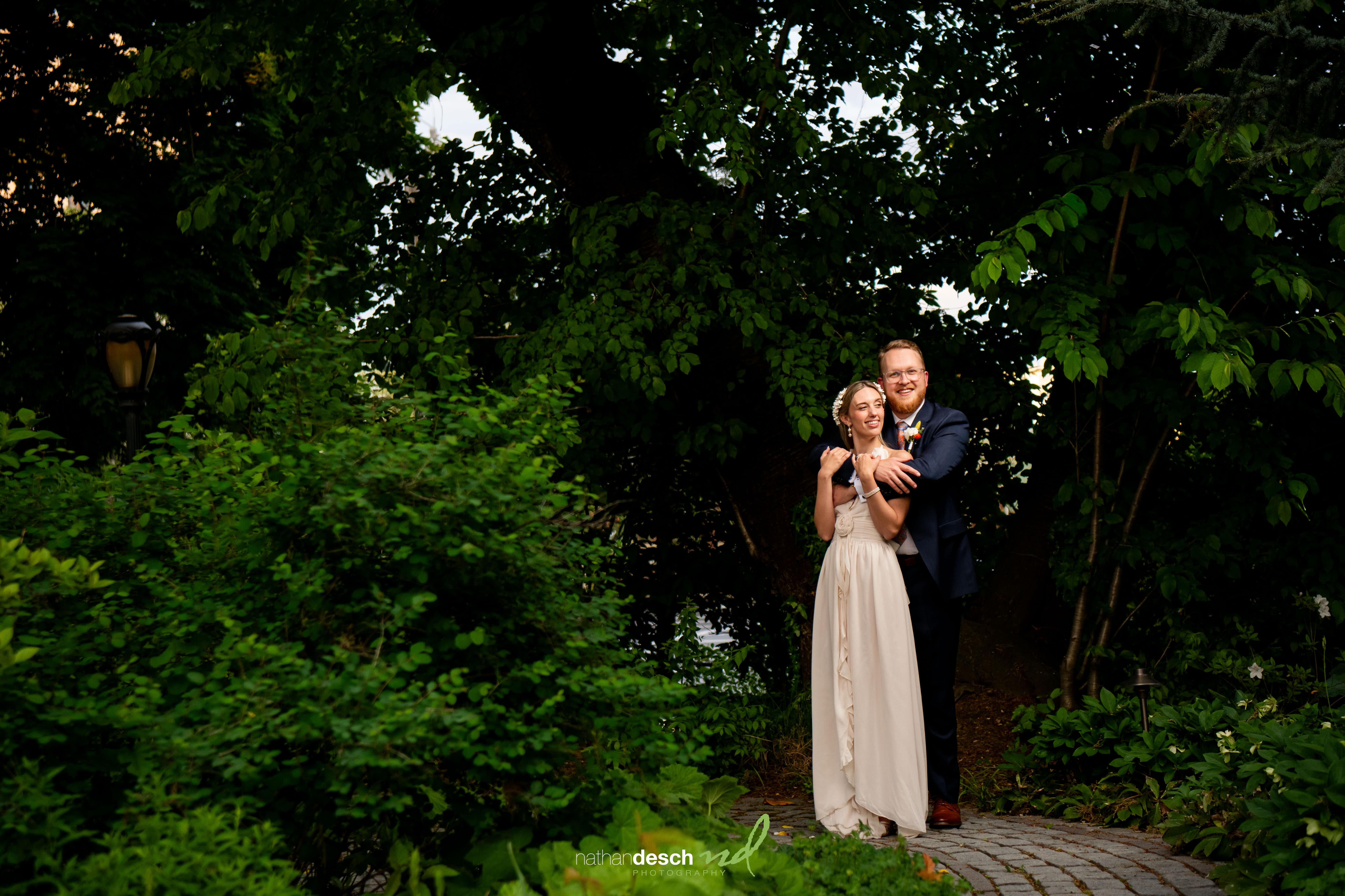 Bride and groom smiling and embracing on a cobblestone path with lush greenery at University and Whist Club wedding.