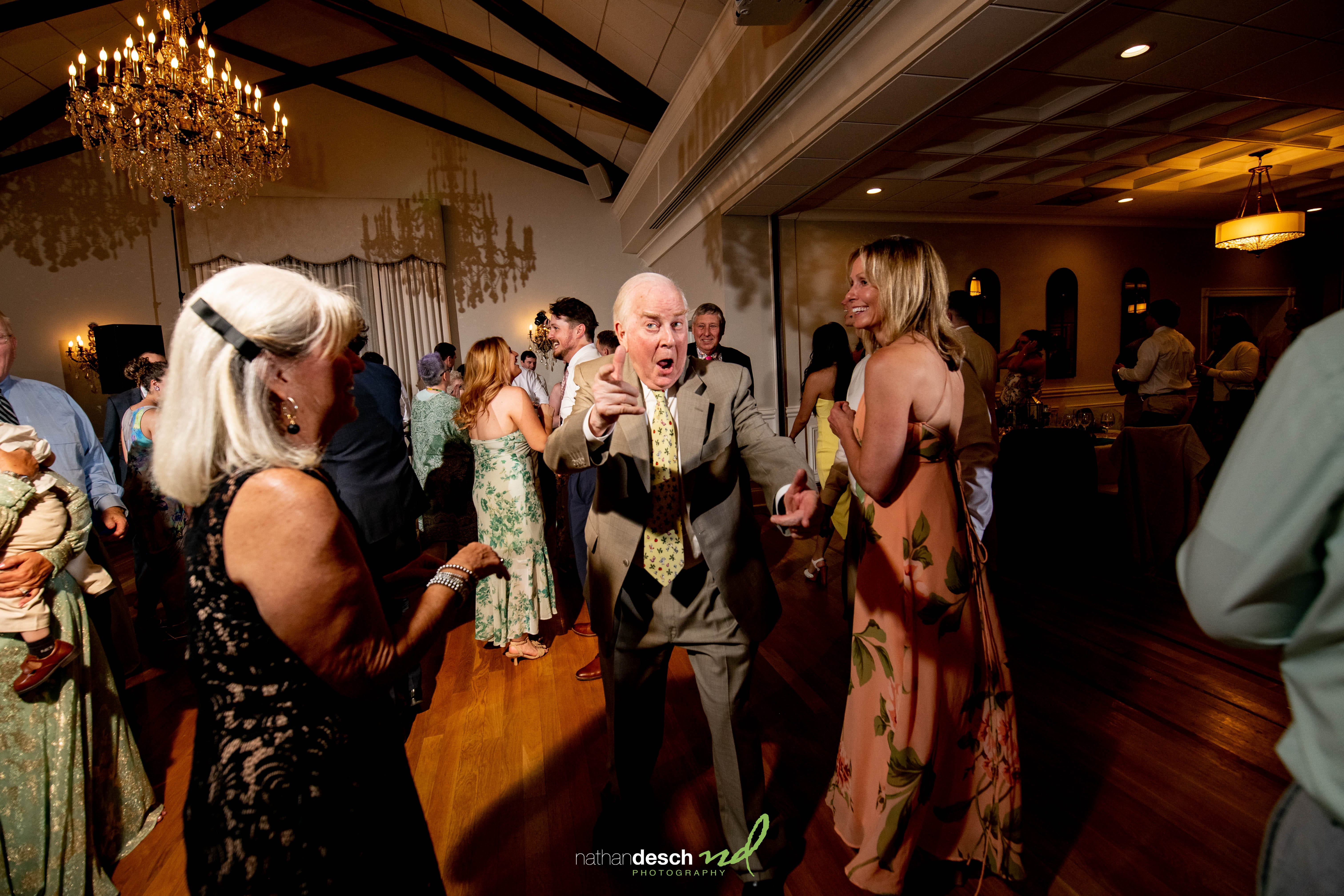 Older man in gray suit dancing energetically at University and Whist Club wedding, guests celebrating under sparkling chandelier.