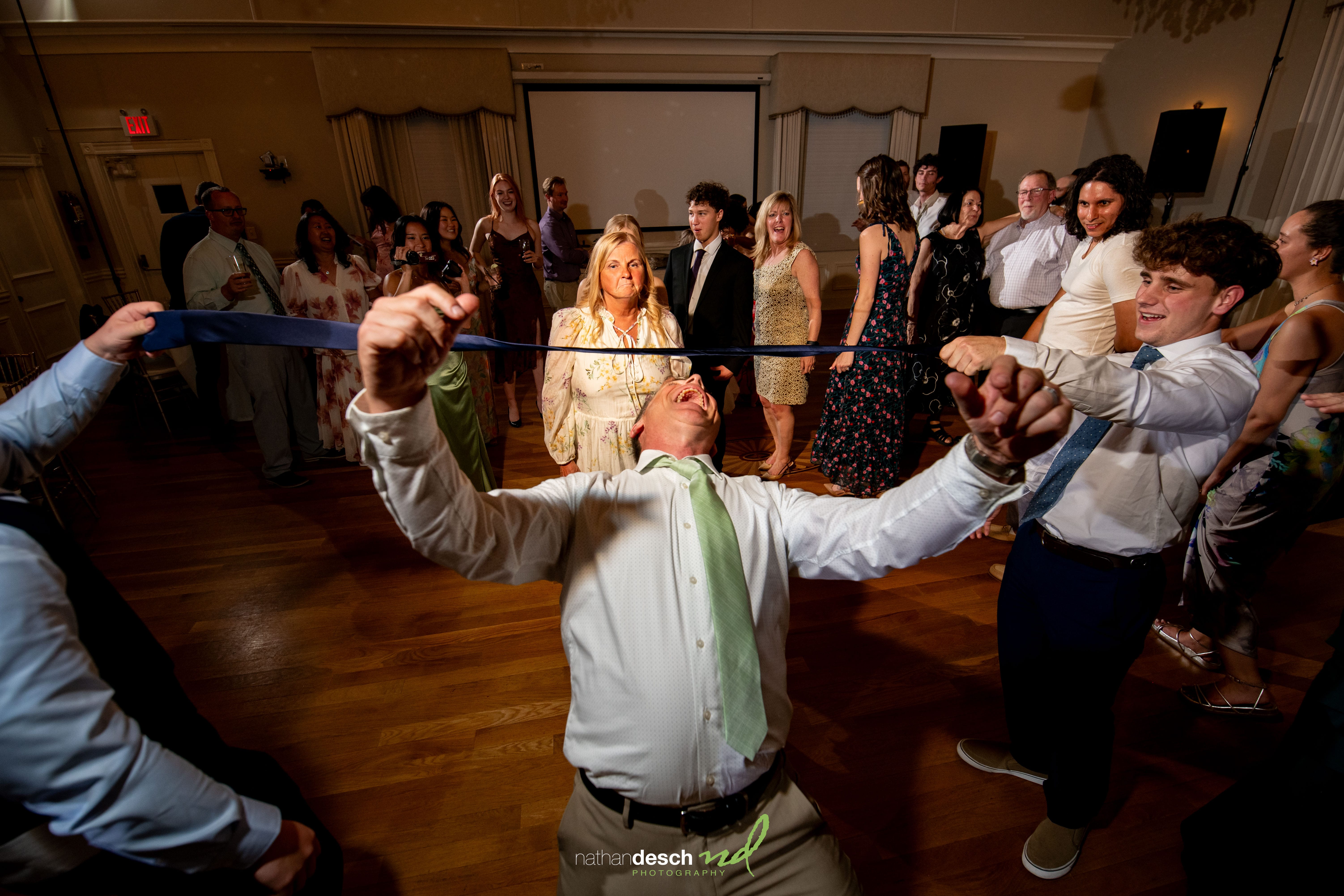 Guests in formal attire smile and watch a man lean back under a blue tie at Garrett and Ashley’s ballroom wedding reception.