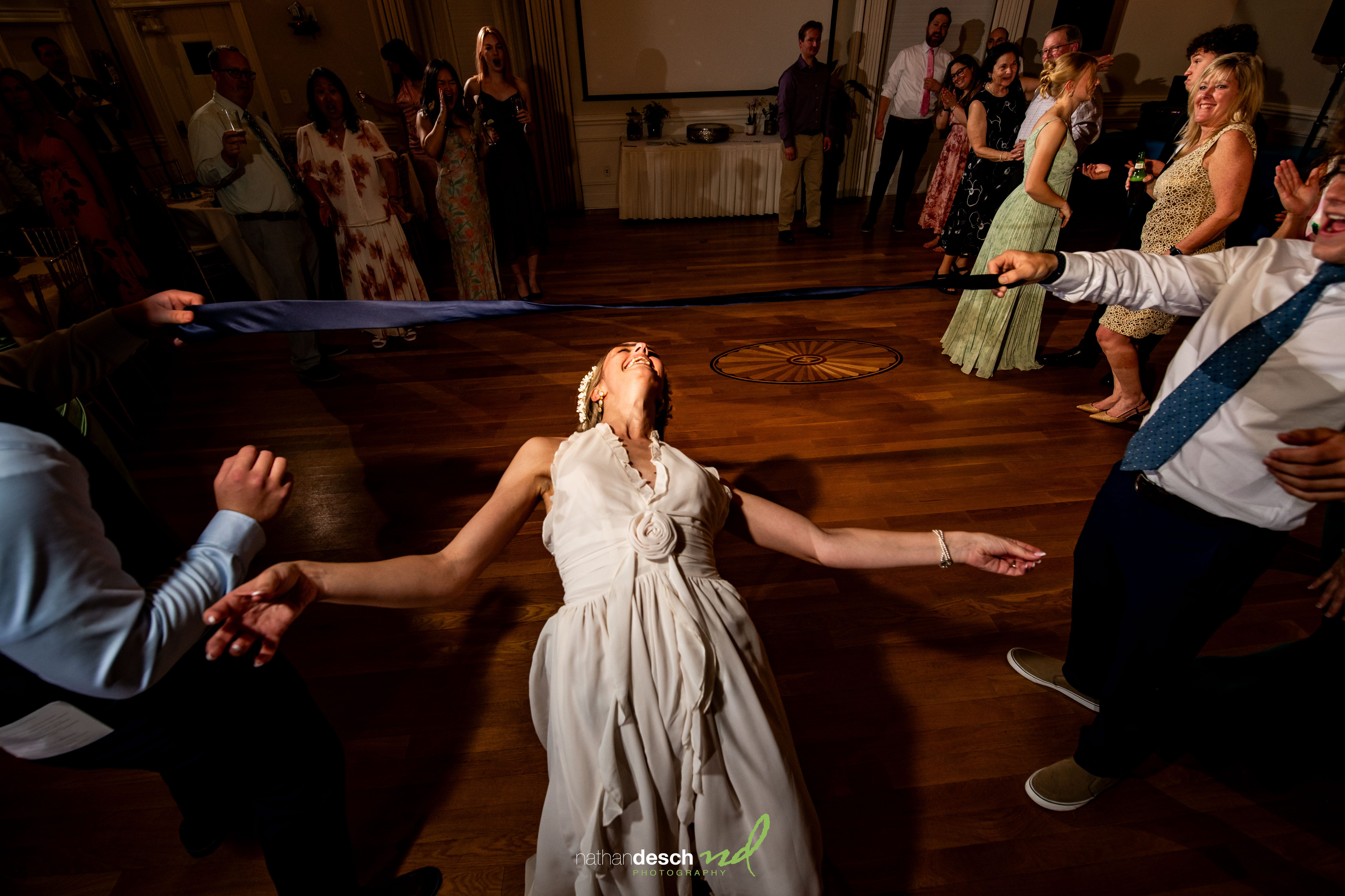 Bride Ashley in white dress does the limbo at joyful University and Whist Club wedding reception as guests smile and watch.