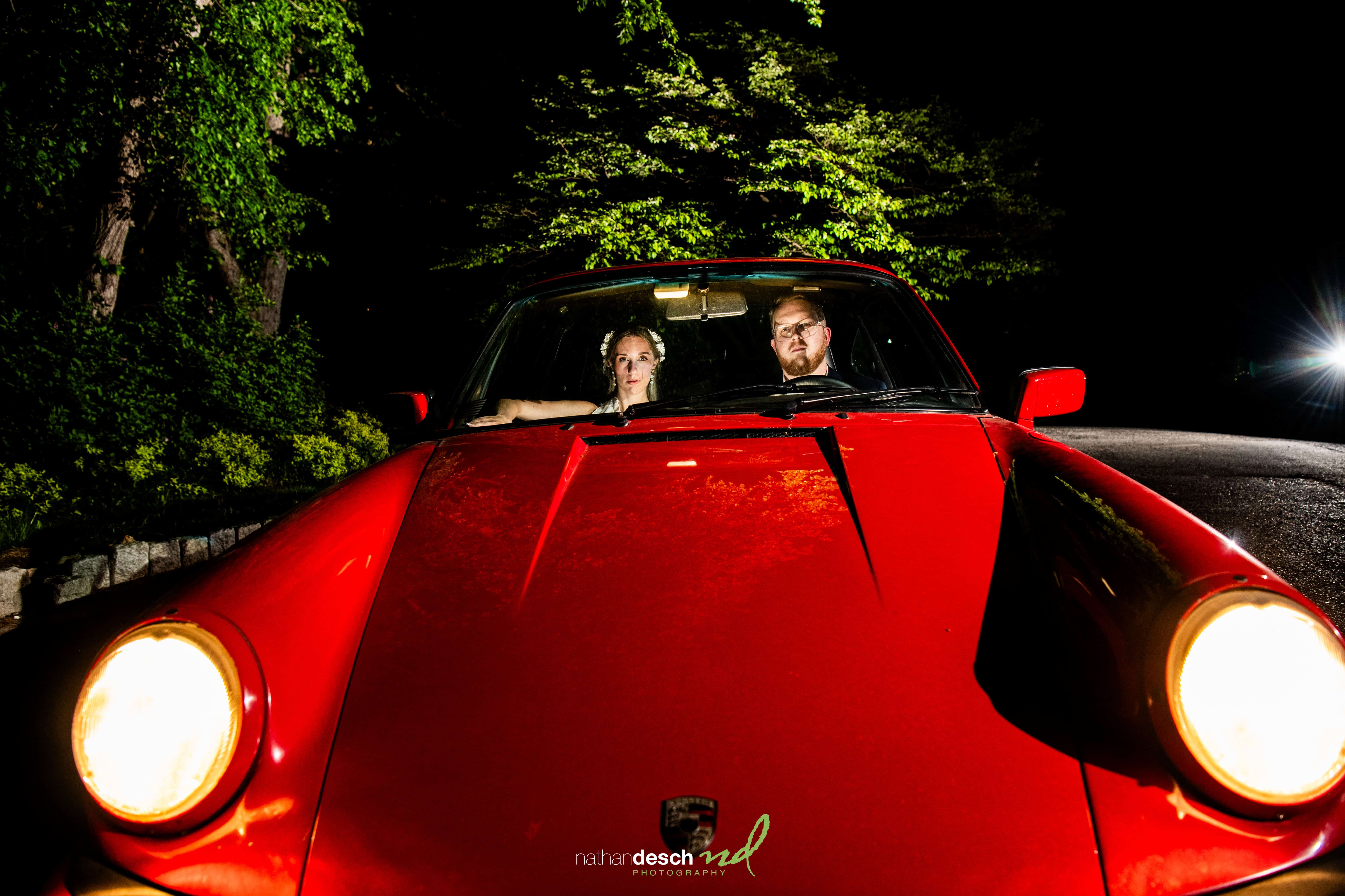 Newlyweds Garrett and Ashley in a red Porsche at night after their University and Whist Club wedding, headlights glowing.