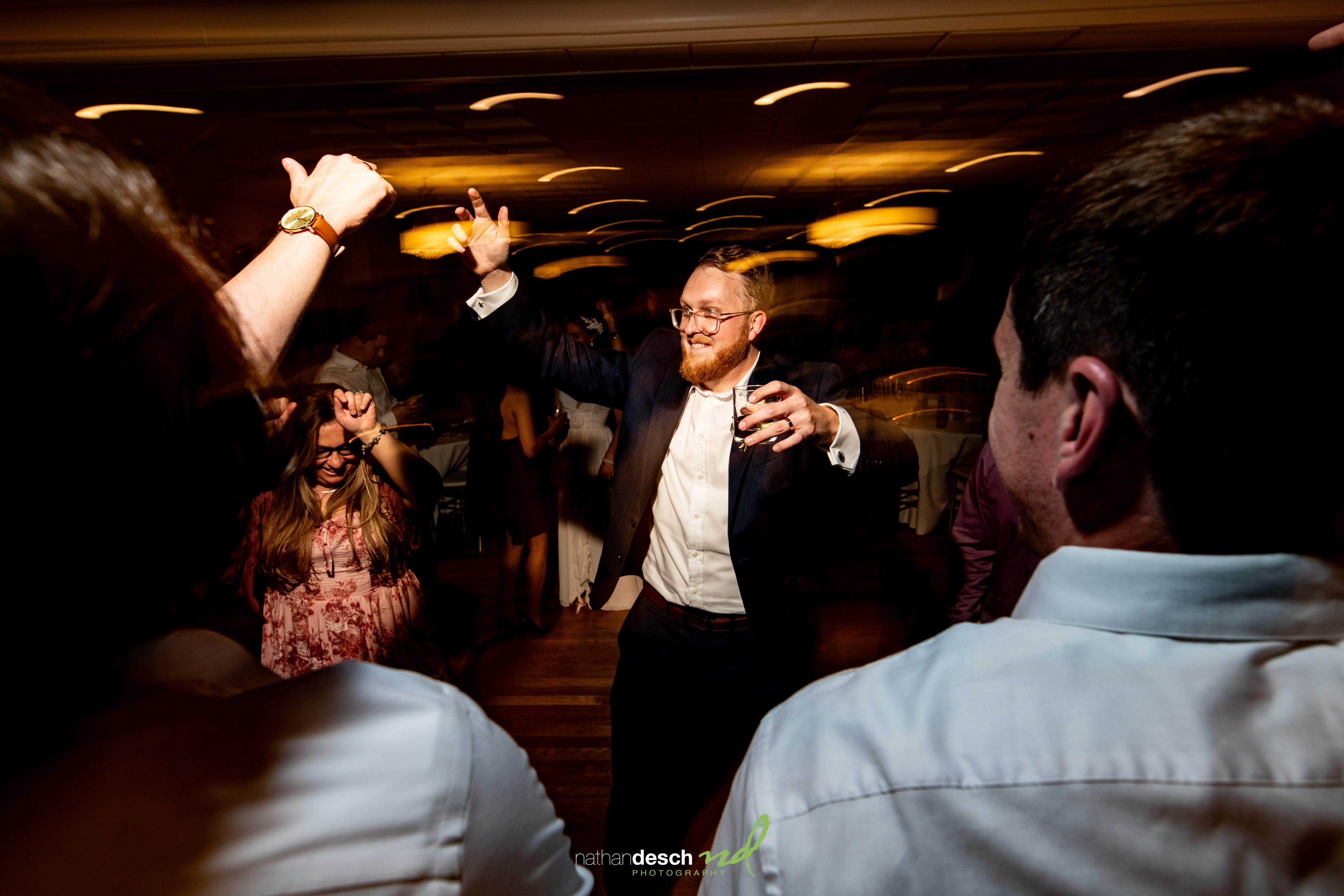 Wedding guest in suit dancing energetically at Garrett and Ashley's University and Whist Club reception, blurred lights overhead.