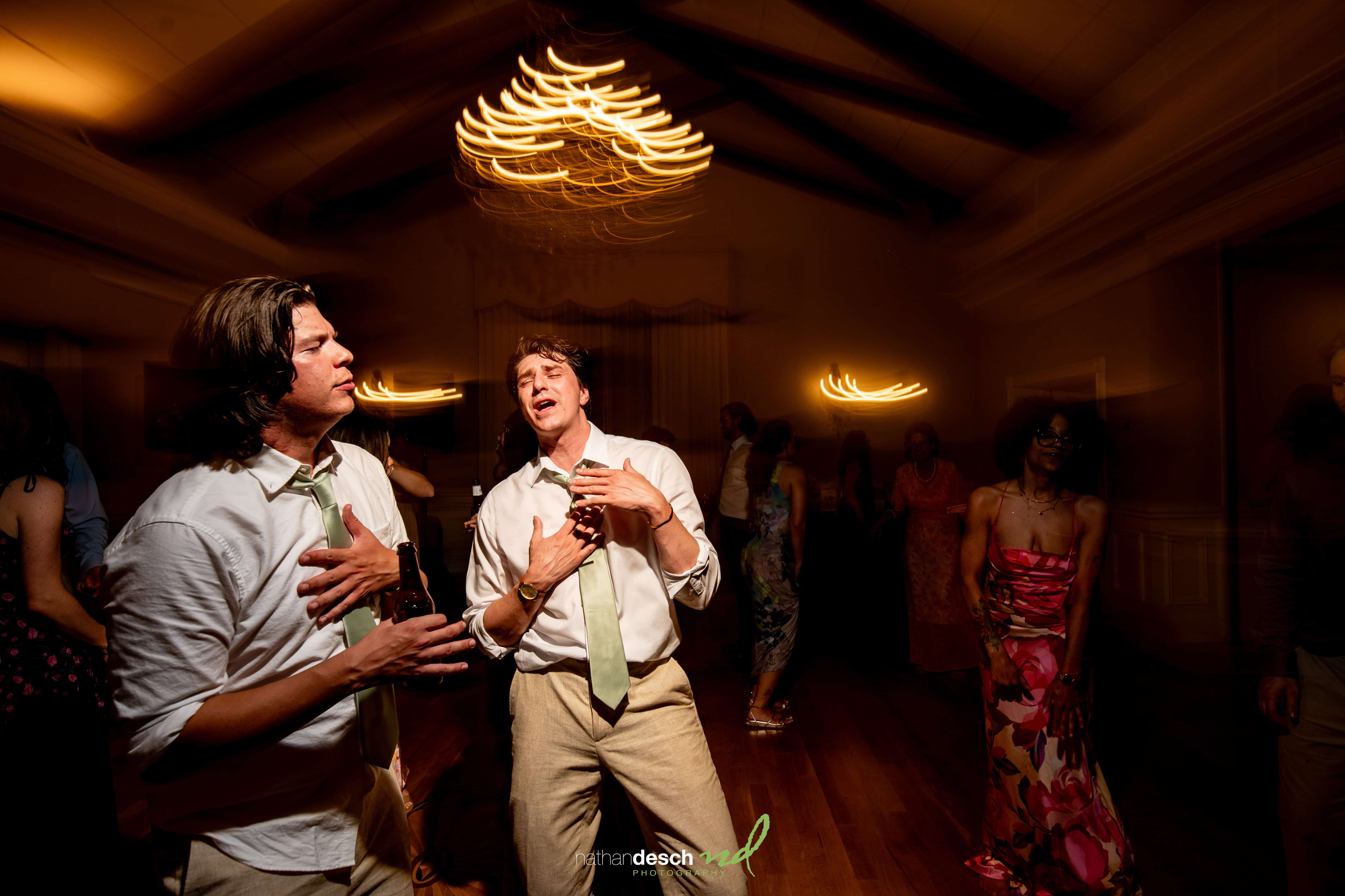 Groom and guest dance and sing at lively University and Whist Club wedding, warm lights creating energetic party atmosphere.