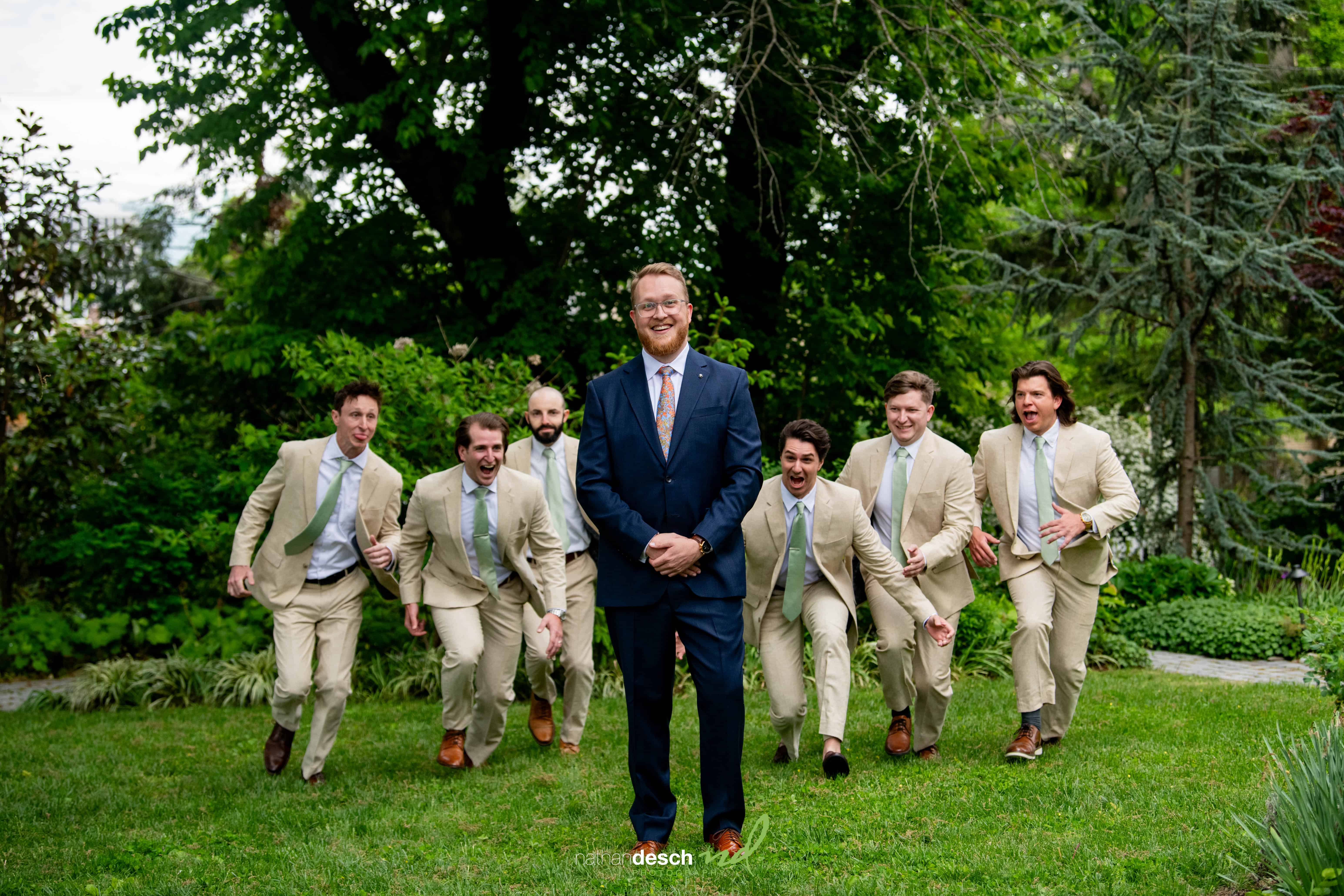 Groom in navy suit smiles as groomsmen in tan suits run toward him in lush garden at University and Whist Club wedding.