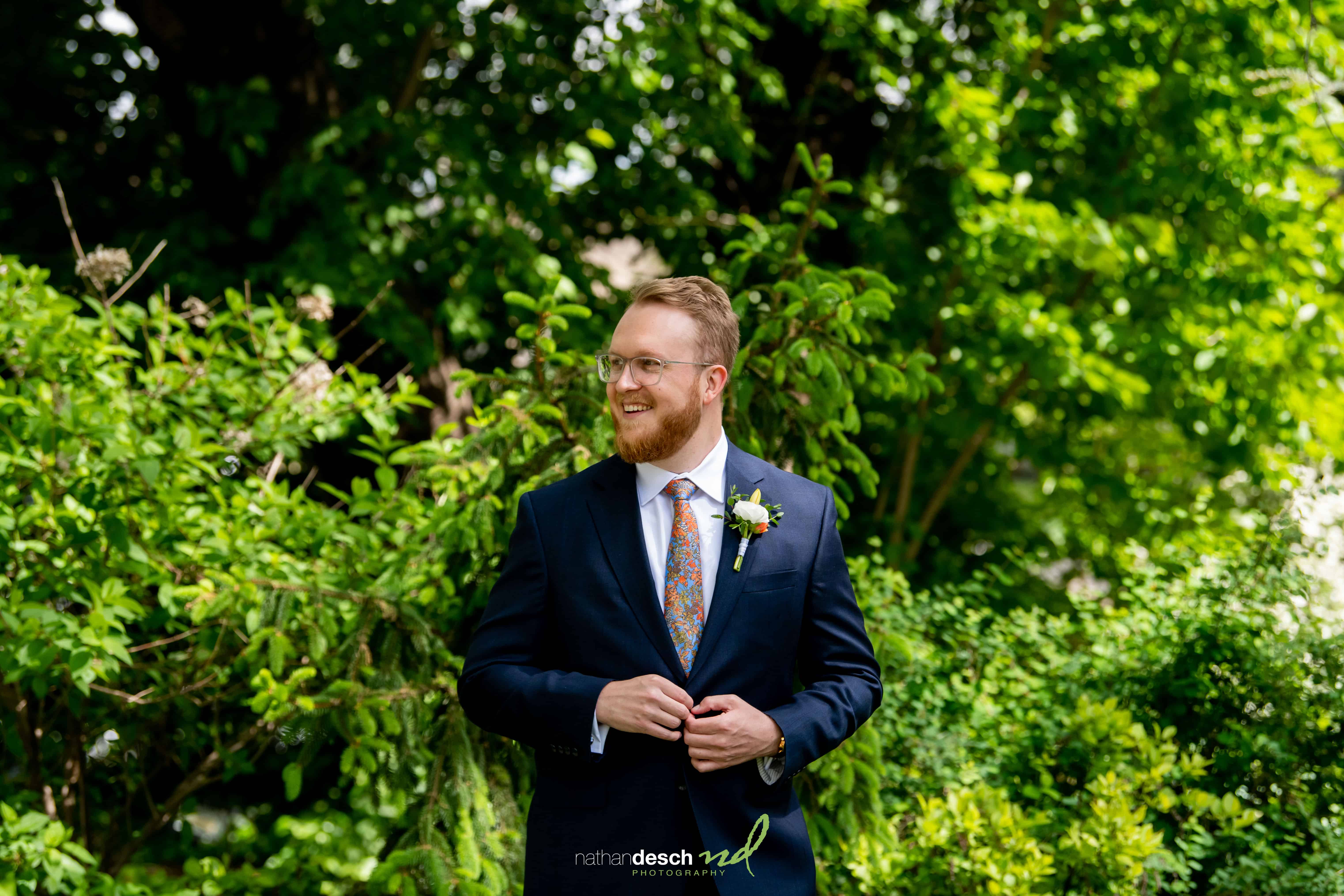Groomsman in dark suit with floral tie smiles before lush greenery at Garrett and Ashley's University and Whist Club wedding.