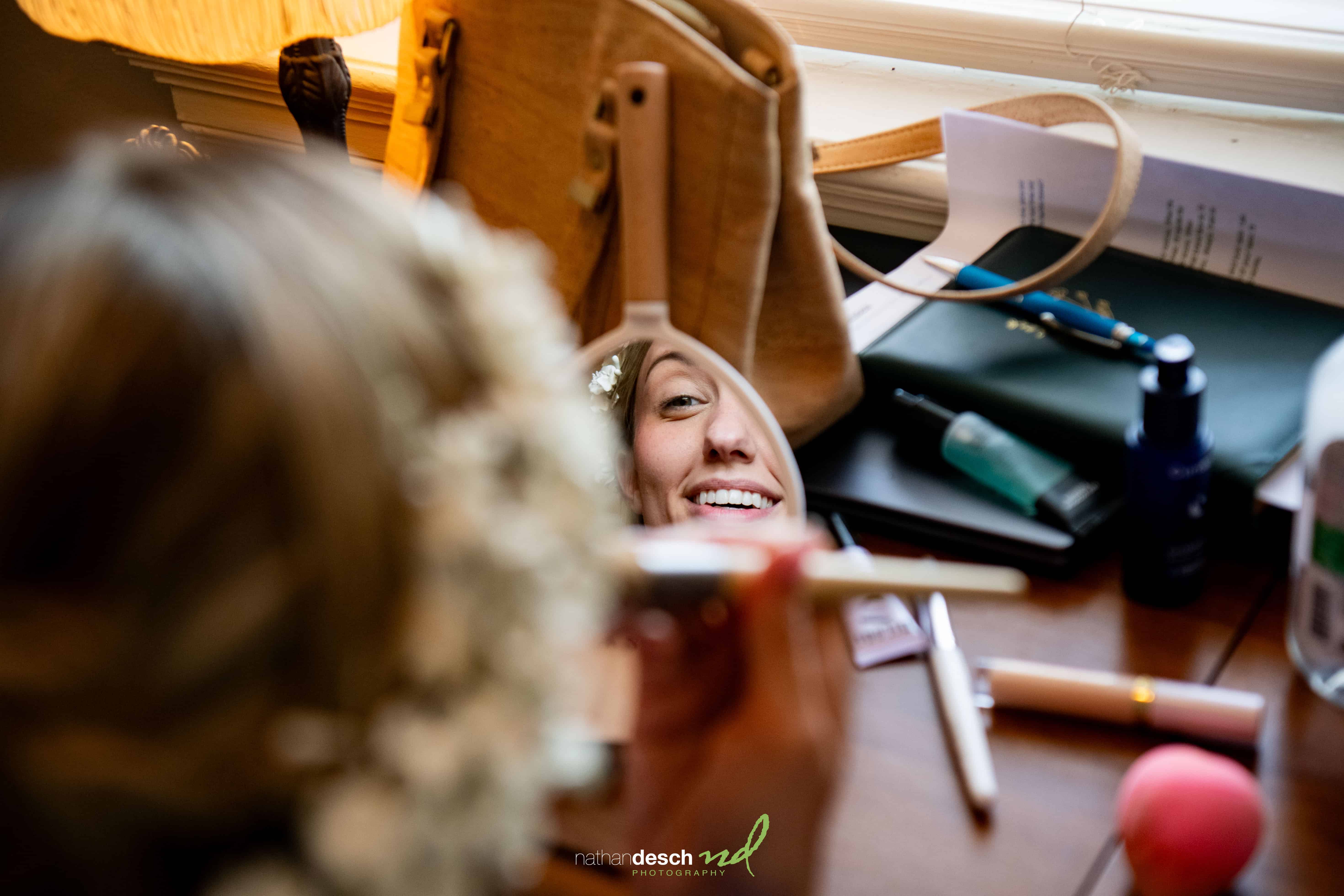 Bride with floral hairpiece smiles in mirror while doing makeup at cluttered table before Garrett and Ashley's wedding.