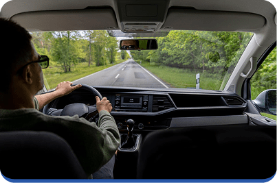 A person wearing glasses is driving a car down an empty, tree-lined road on a bright day, as seen from the passenger seat. The dashboard and steering wheel are visible in the foreground.