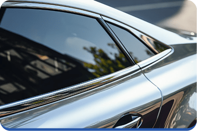 A close-up view of the rear side window and door of a shiny silver car, reflecting nearby buildings and greenery. The photo highlights the car’s sleek lines and smooth metallic surface.