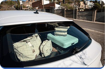 A view through the rear window of a white car shows several bags and a suitcase packed in the trunk. Houses and a quiet street are visible in the background.