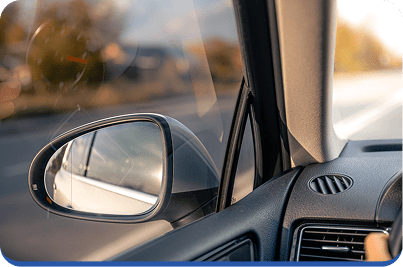 Close-up view from inside a car, showing the side mirror, partially open window, and a hand on the steering wheel, with a blurred road and landscape visible outside.