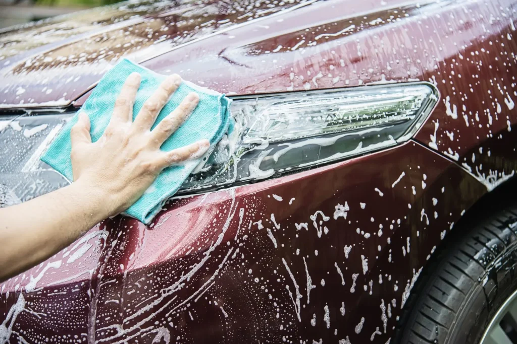 A person’s hand uses a blue microfiber cloth to wash the headlight area of a red car, which is covered in soap suds and water.