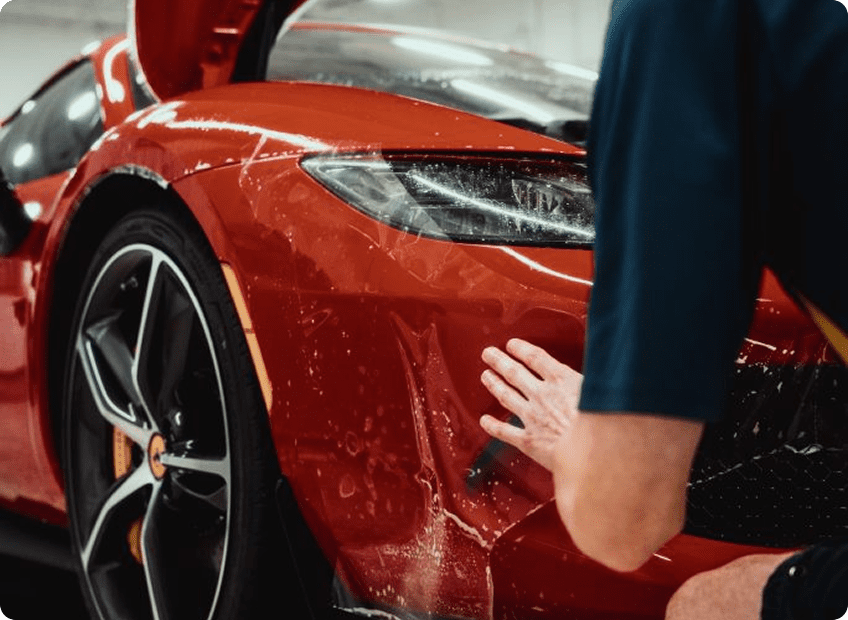 A person applies a protective film to the front bumper of a shiny red sports car, with the cars hood open and water droplets visible on the surface.