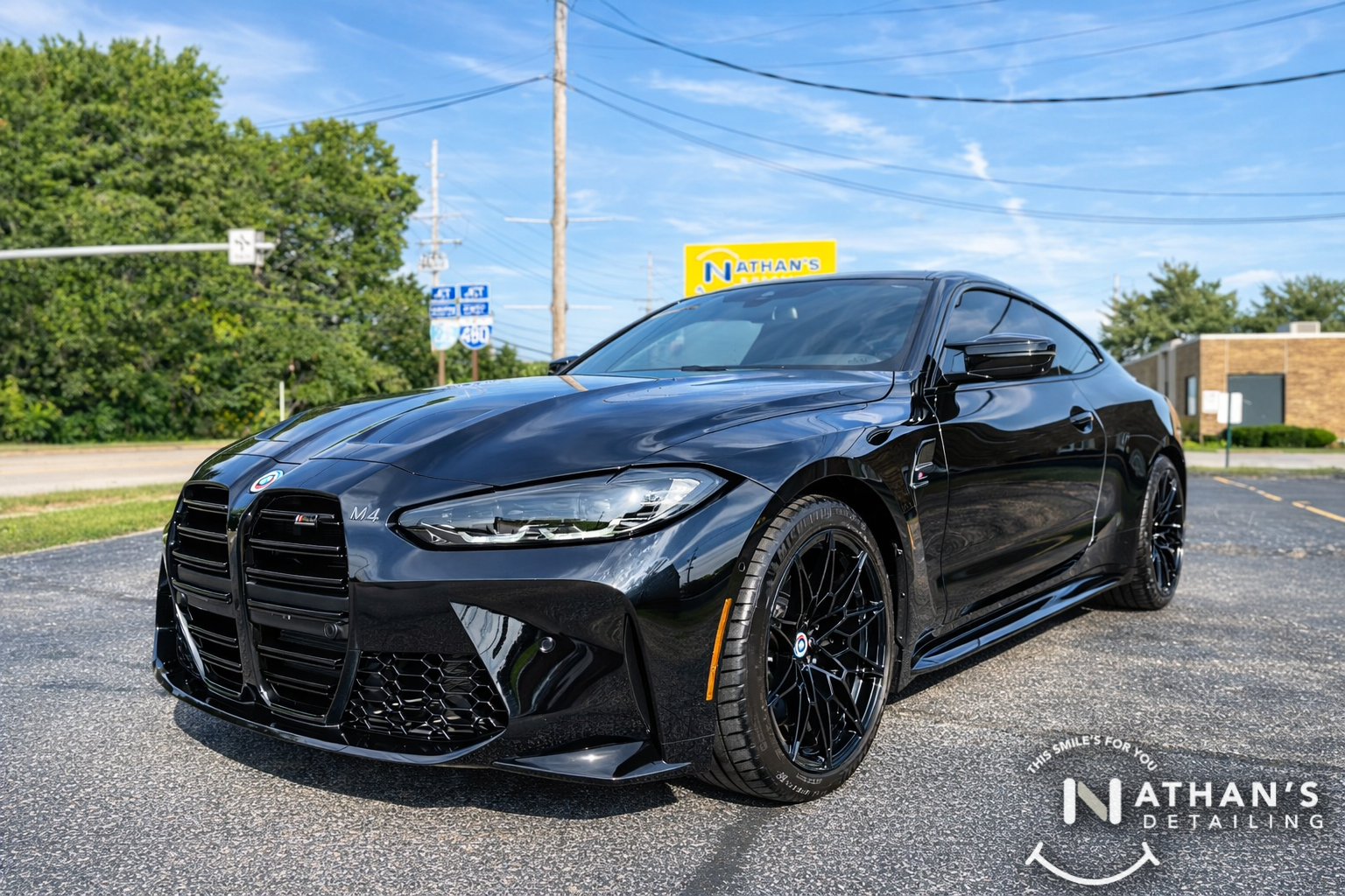 A shiny black BMW M4 coupe is parked in a lot on a sunny day, reflecting blue sky and clouds. The car is clean, with Nathan’s Detailing signage in the background.