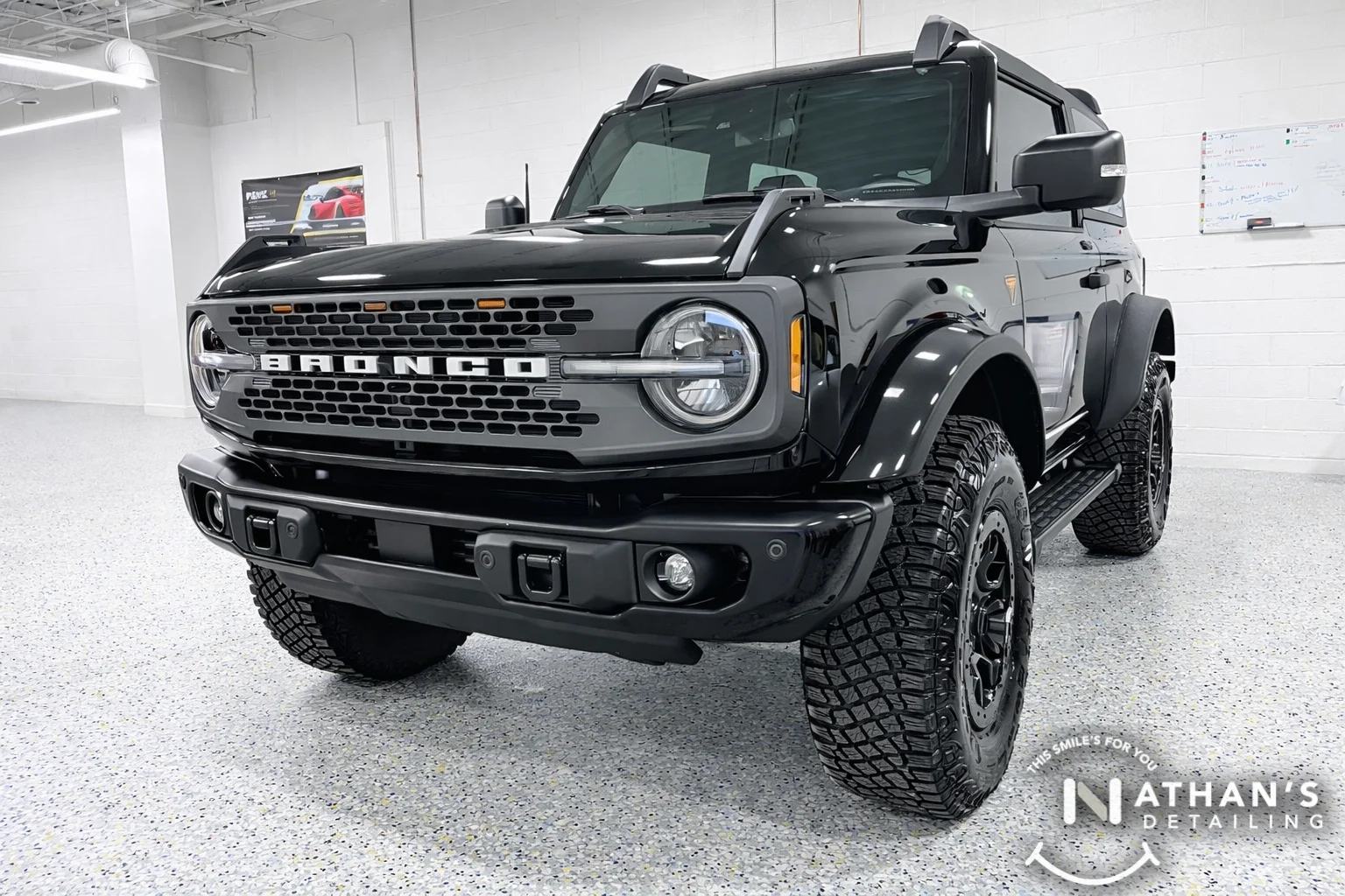 A shiny black Ford Bronco SUV with large off-road tires is parked indoors on a speckled floor. The front grill displays the BRONCO logo, and the vehicle appears newly detailed. “Nathan’s Detailing” logo is in the corner.