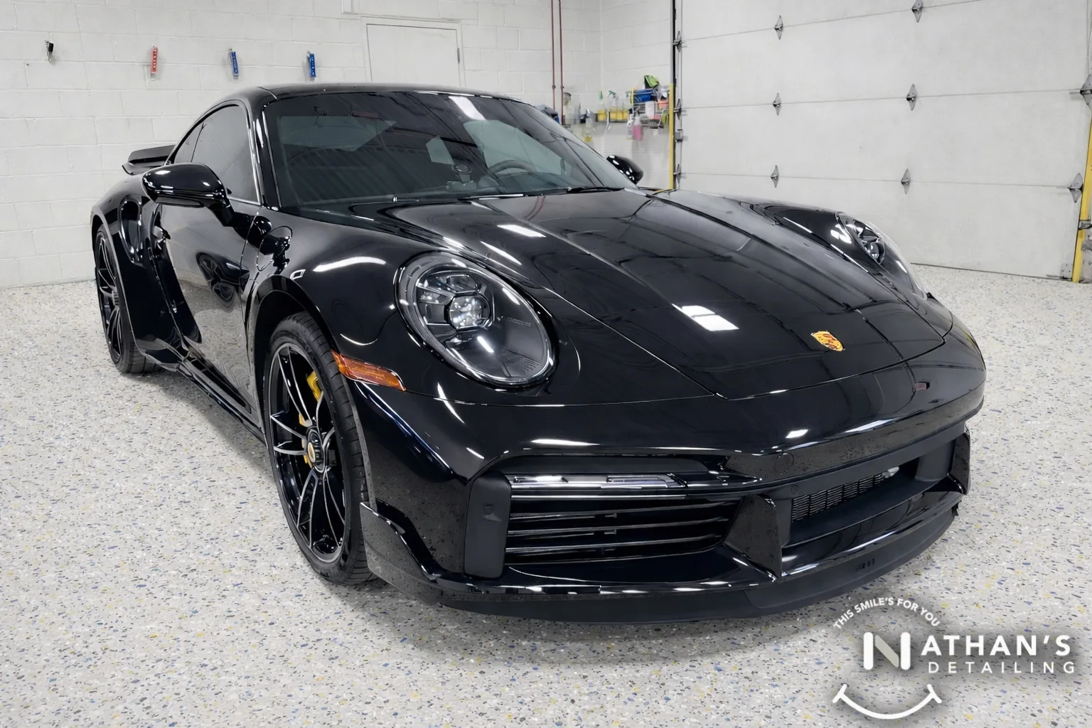 A shiny black Porsche sports car is parked inside a clean, well-lit garage with speckled flooring. The cars exterior is highly polished and reflects the surroundings. Nathans Detailing logo is visible in the bottom right corner.