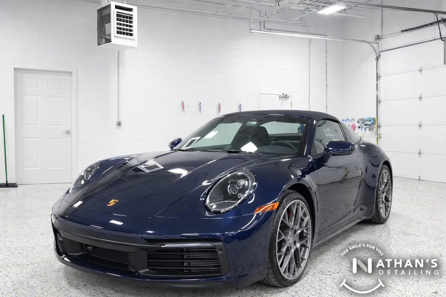A dark blue Porsche sports car is parked inside a clean, well-lit garage with white walls and a speckled floor. Car detailing tools are hung on the back wall. The Nathans Detailing logo is visible in the lower right corner.
