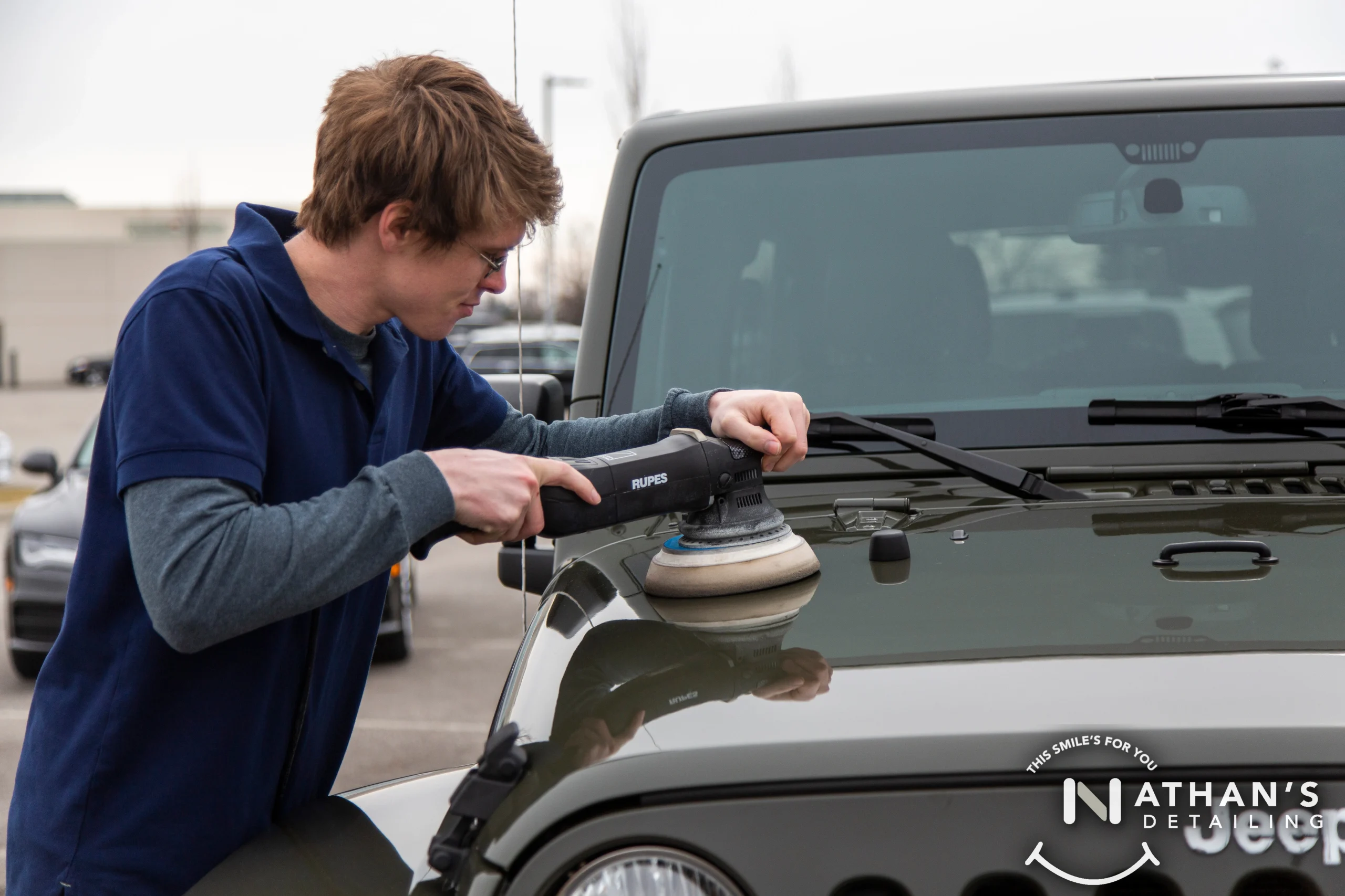 A man wearing glasses uses a polishing machine on the hood of a green Jeep in a parking lot. The logo Nathans Detailing is visible in the bottom right corner.