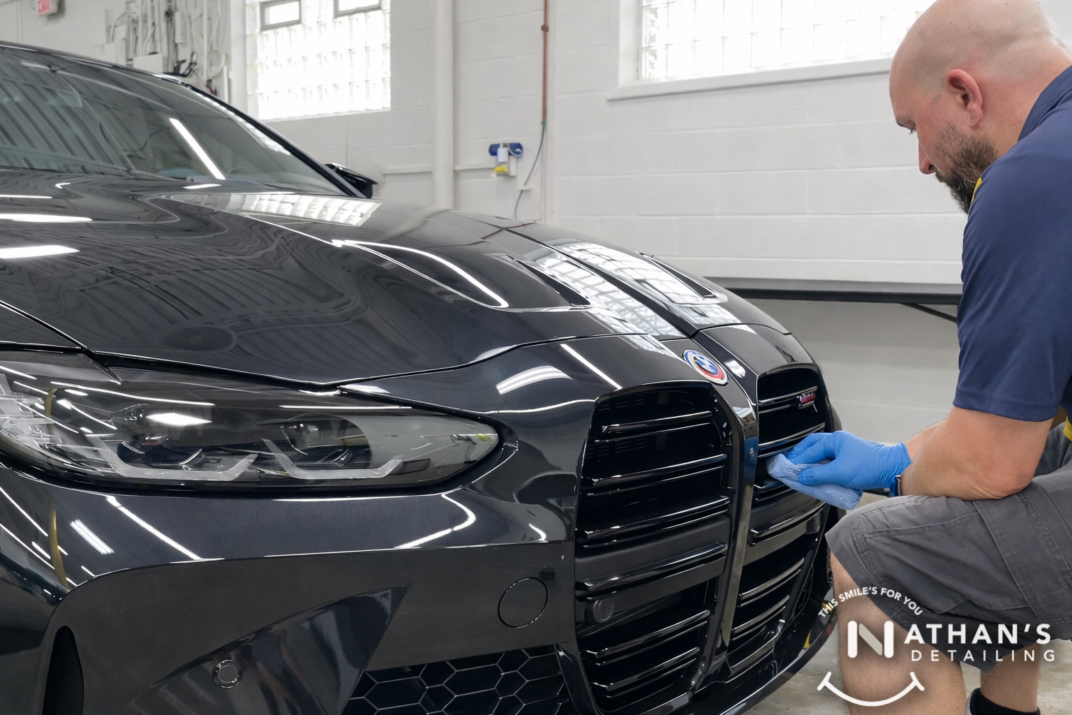 A man wearing gloves uses a tool to clean or detail the black front grille of a shiny BMW car inside a garage. The Nathan’s Detailing logo is visible in the bottom right corner.