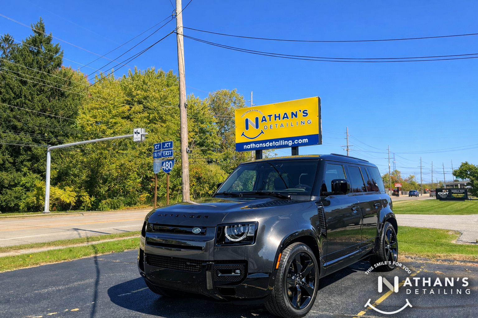 A black Land Rover Defender is parked outside on a sunny day near a road, with a yellow Nathan’s Detailing sign in the background and trees and utility poles visible. Nathan’s Detailing logo is in the corner.