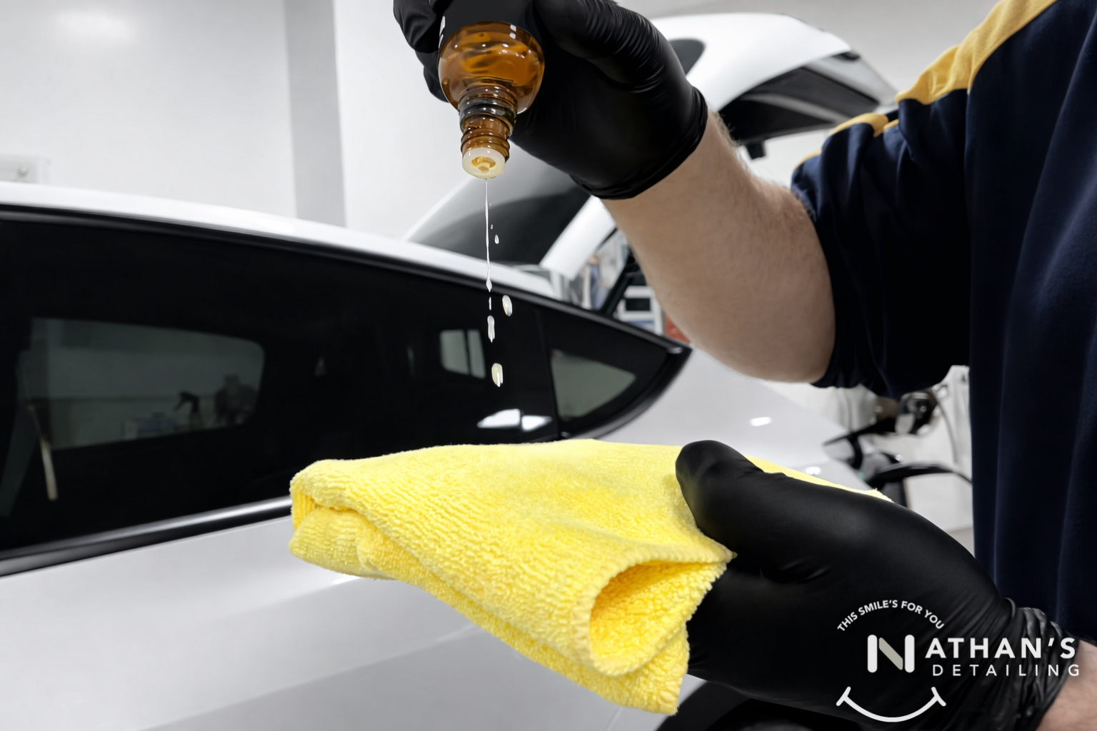 A person wearing black gloves pours liquid onto a yellow microfiber cloth in front of a white car. The words “Nathan’s Detailing” are visible in the bottom right corner.