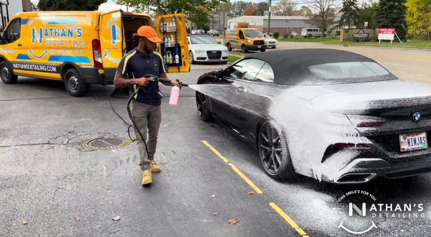 A man sprays foam onto a black convertible BMW during a mobile car wash in a parking lot. A yellow Nathans Detailing van and another car are visible in the background.