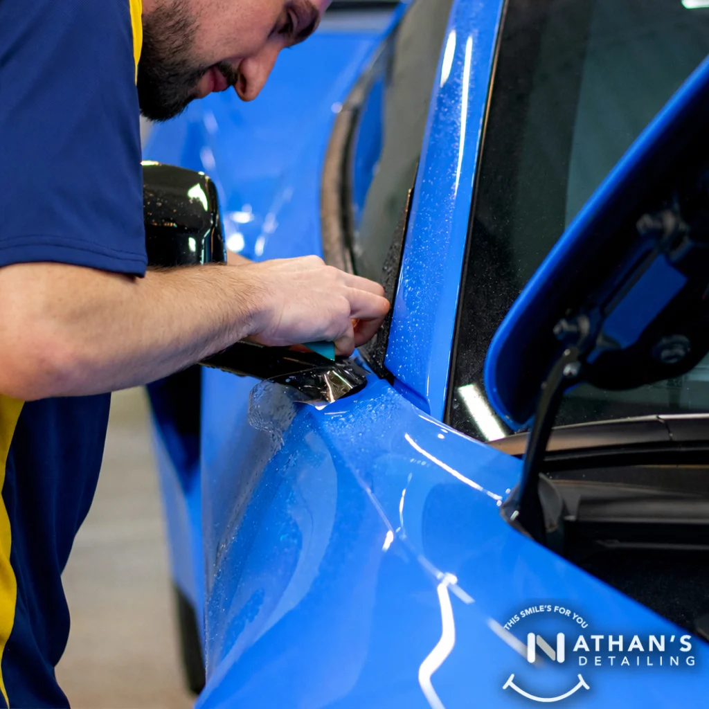 A person applies a protective film to the side mirror of a shiny blue car, focusing intently. The Nathan’s Detailing logo is visible in the bottom right corner.